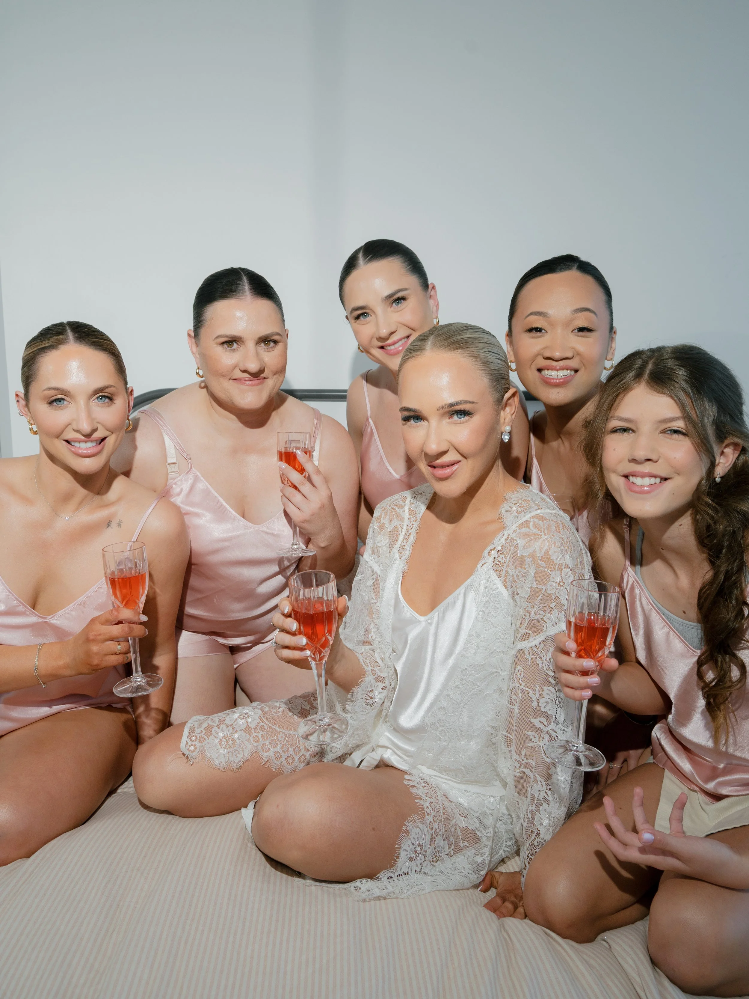 Group of six women in pajamas sitting on a bed, holding glasses of pink drinks, smiling at the camera.