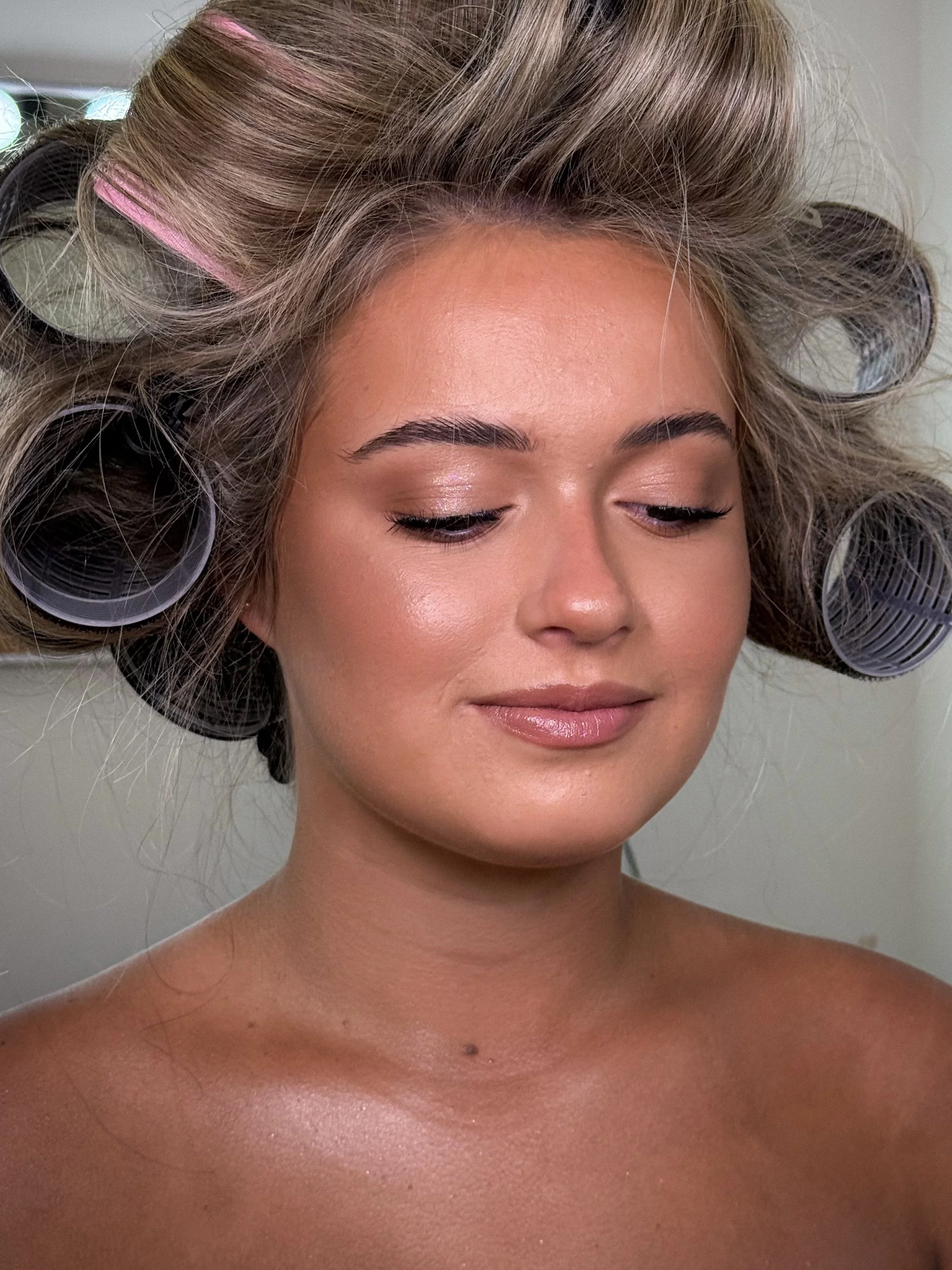 A woman with medium skin tone and blonde hair with gray streaks is getting her hair styled with hair rollers in a salon, her eyes closed and a gentle smile on her face.