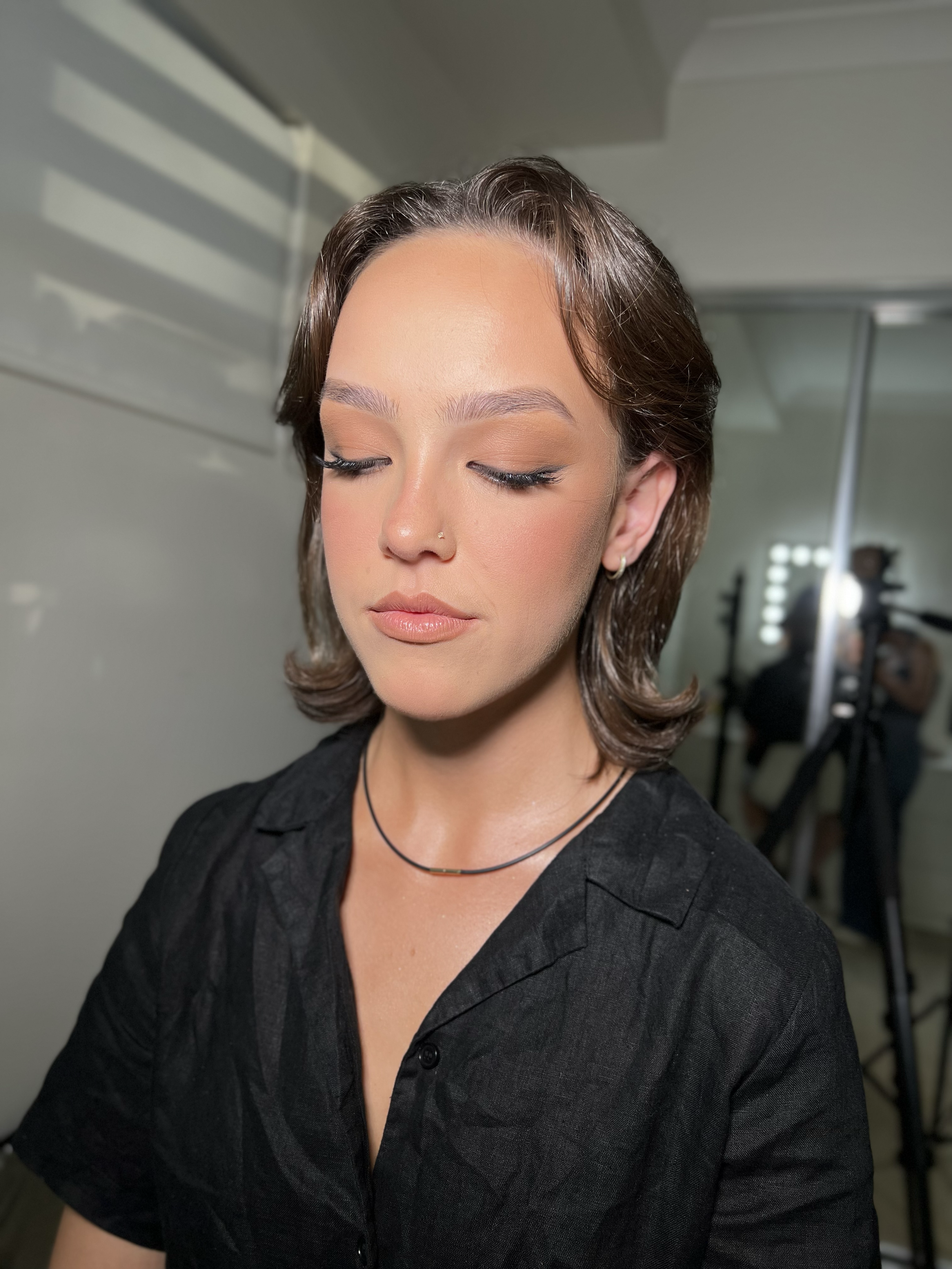Close-up of a woman with short brown hair, light makeup, wearing a black shirt and thin necklaces, with her eyes closed, in a professional photo studio setting.