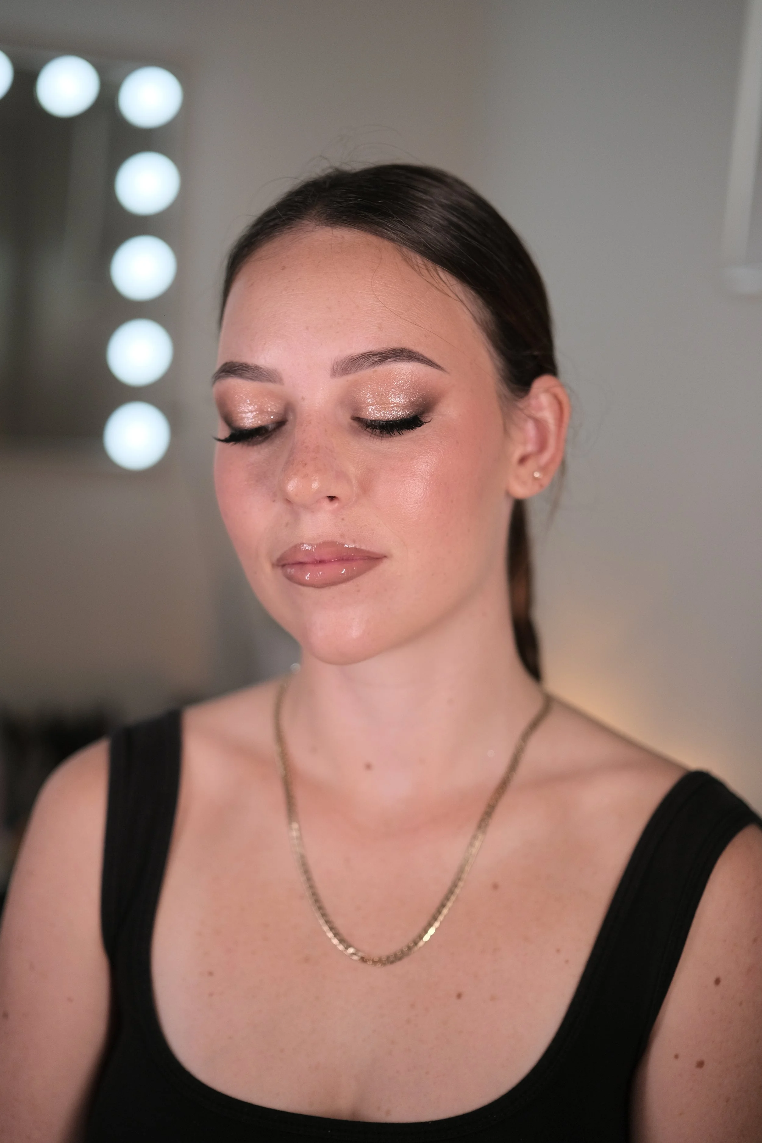 A woman with closed eyes displaying a makeup look with shimmery eyeshadow, glossy lips, and a sleek hairstyle, standing in front of a mirror with round lights.