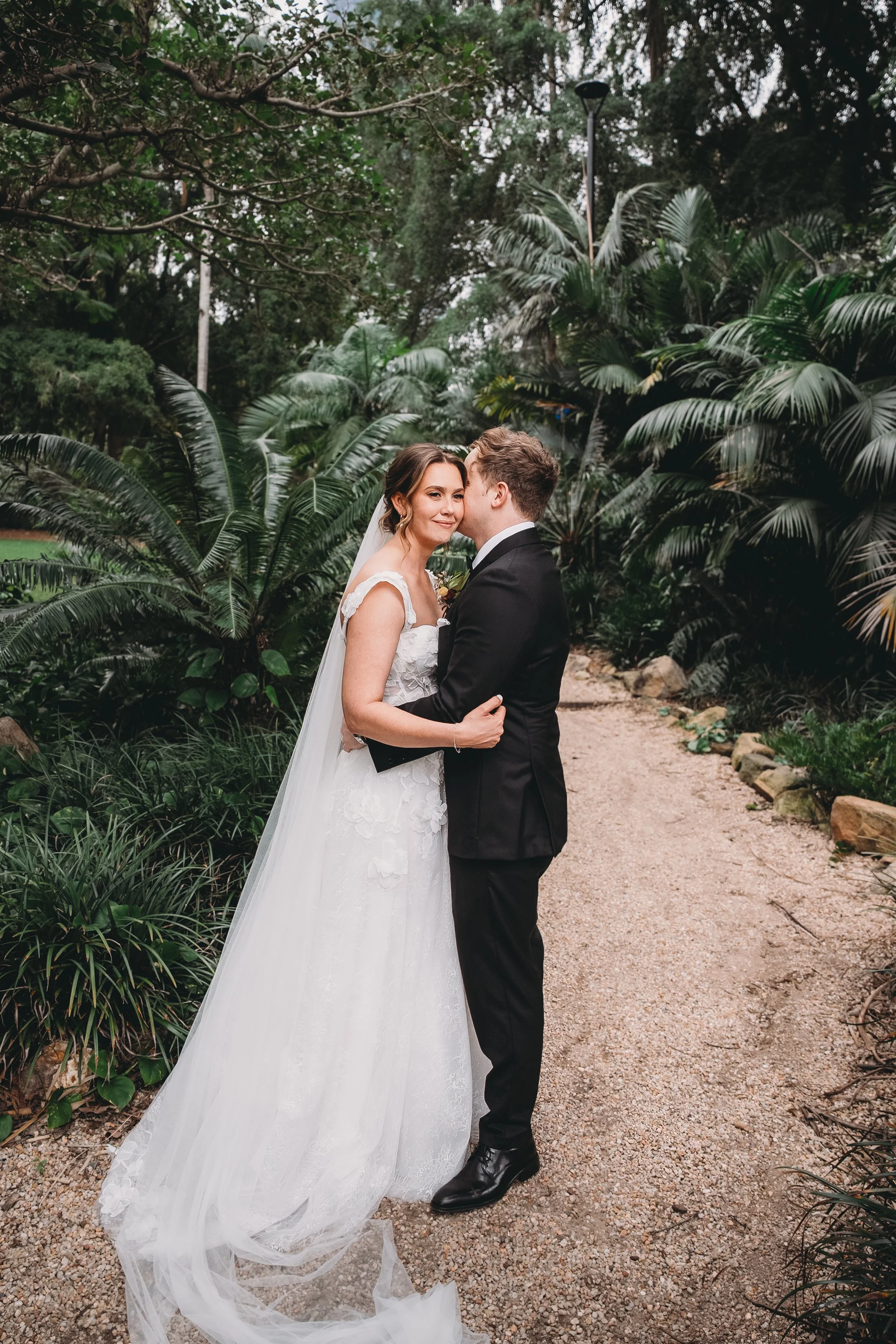 A bride and groom embrace on a garden path surrounded by lush green plants, with the groom kissing the bride's cheek.
