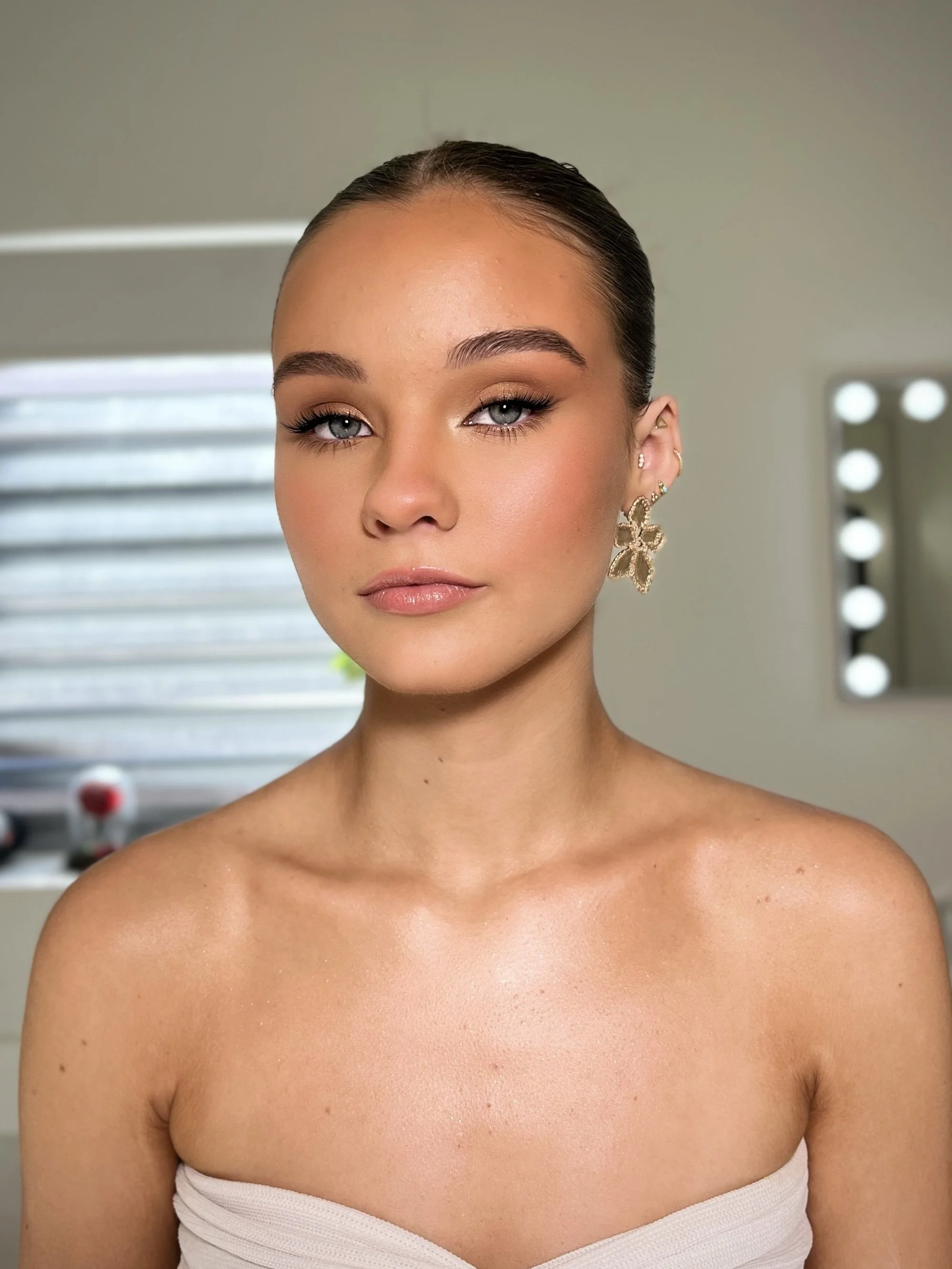 Portrait of a young woman with slicked-back hair, blue eyes, and makeup, wearing large floral earrings, in a room with a mirror and bright lighting.