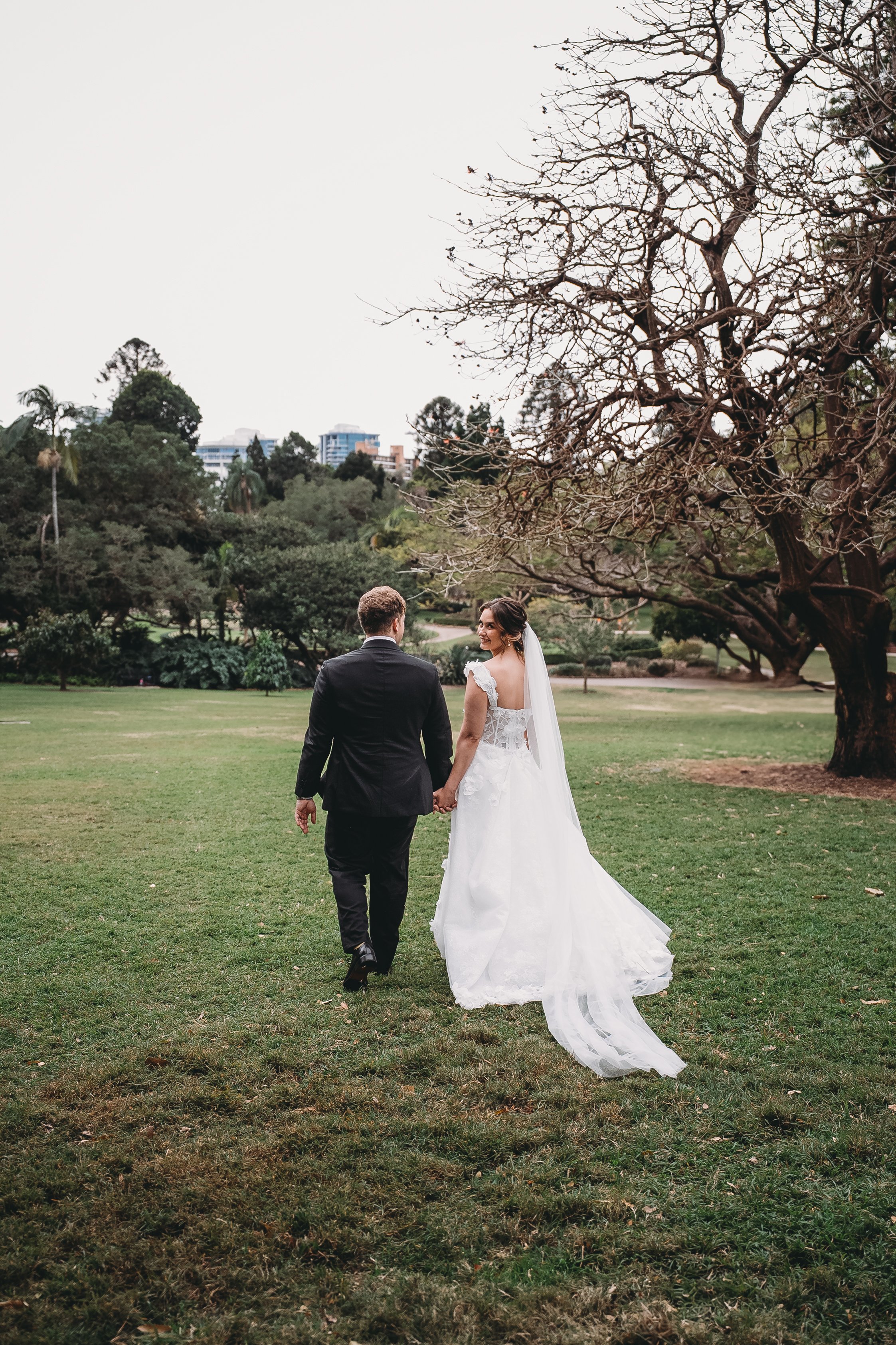 A bride and groom walking hand in hand across a grassy park, with trees and distant city buildings in the background.