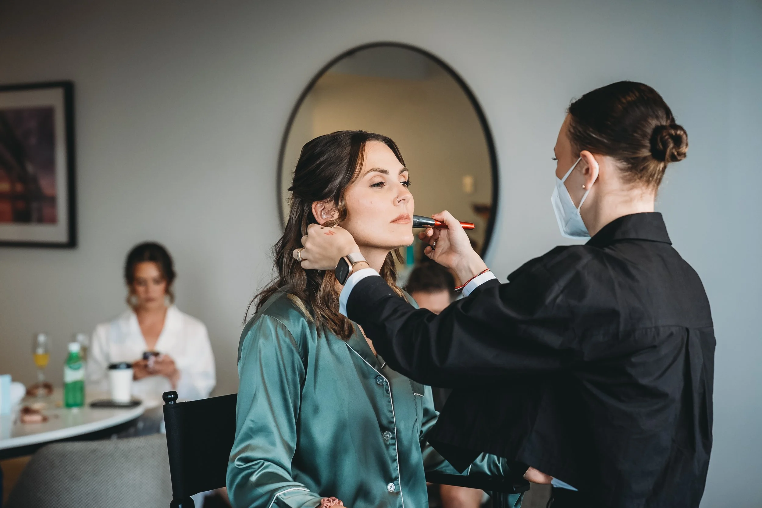 A woman in silk pajamas gets her makeup done by a makeup artist wearing a face mask, in a room with a mirror, table, and other women in the background.