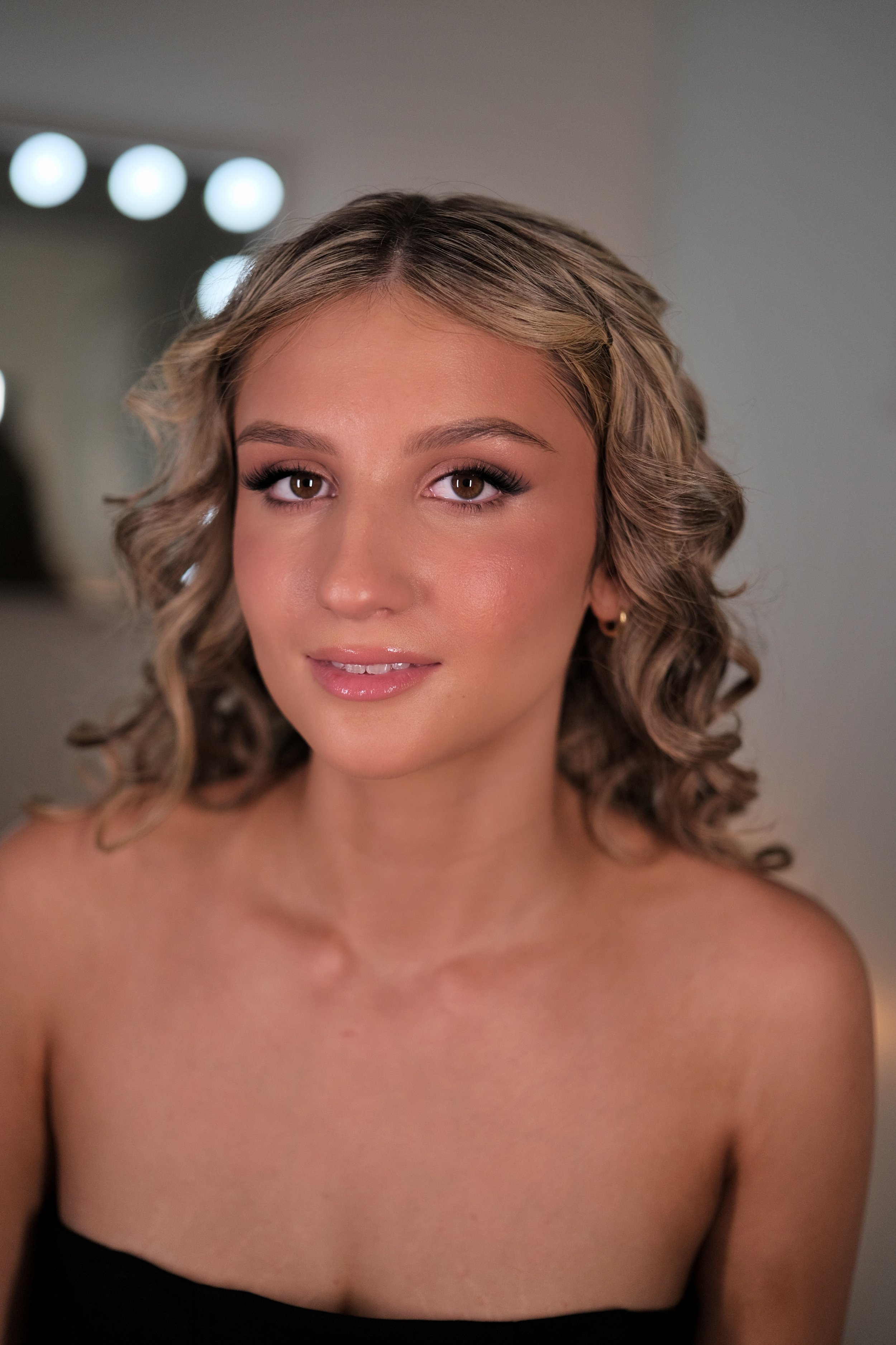 A young woman with shoulder-length curly blonde hair, wearing a black strapless top, smiling softly with makeup including eyeshadow, eyeliner, mascara, and nude lipstick. She is in a room with blurry lights in the background.