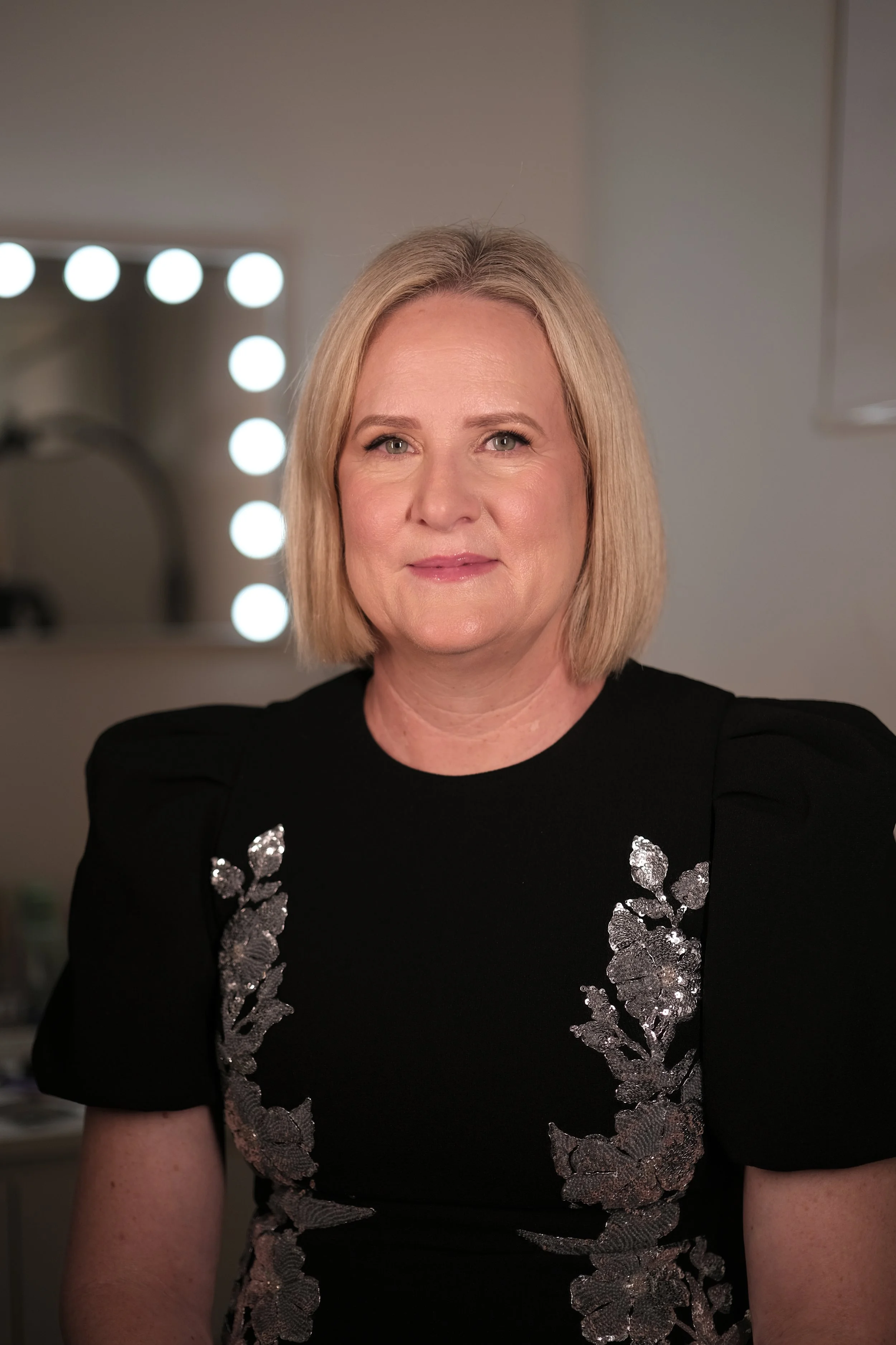 A woman with blonde hair wearing a black dress with silver embroidery, in front of a mirror with bright lights.
