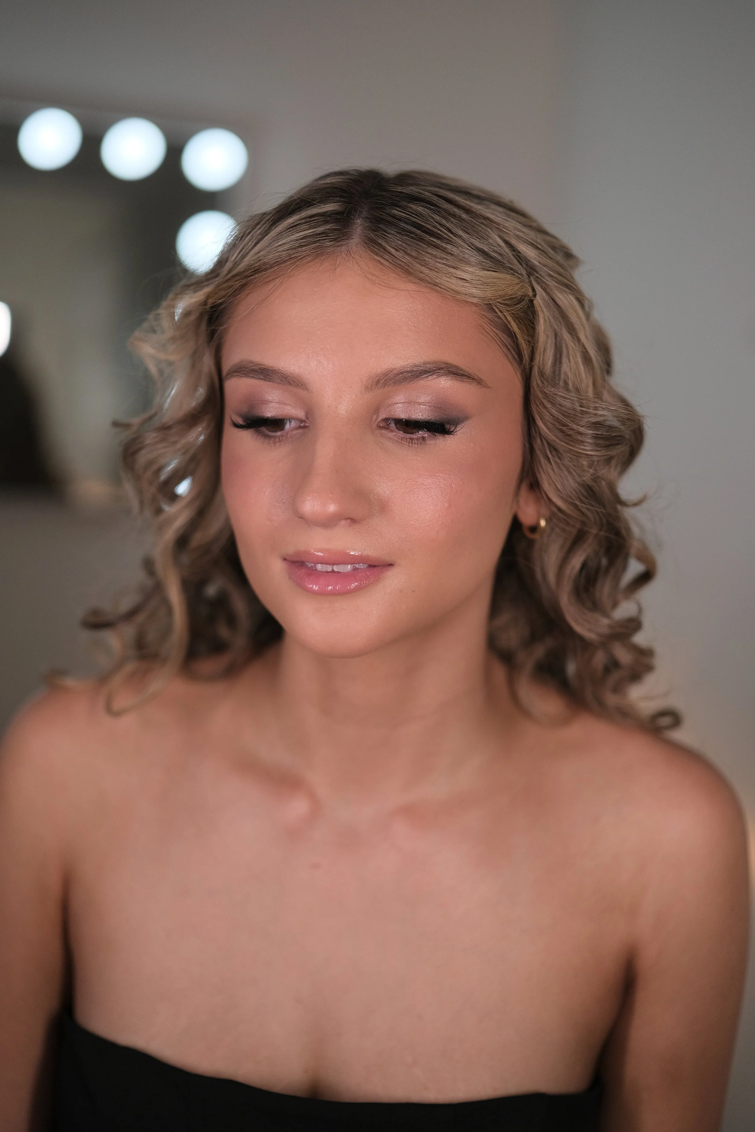 A young woman with curly hair and light makeup looking down, wearing a strapless black top and small earrings.