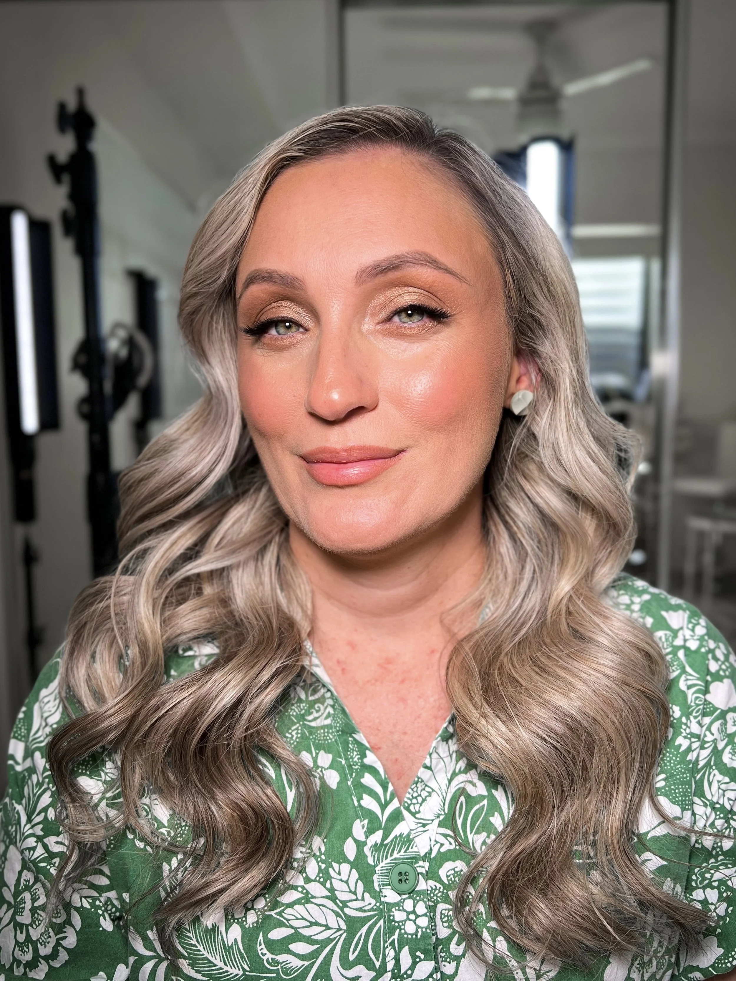 A woman with wavy, blonde hair, wearing makeup, earrings, and a green patterned shirt, smiling in a well-lit room with a mirror and workout equipment in the background.