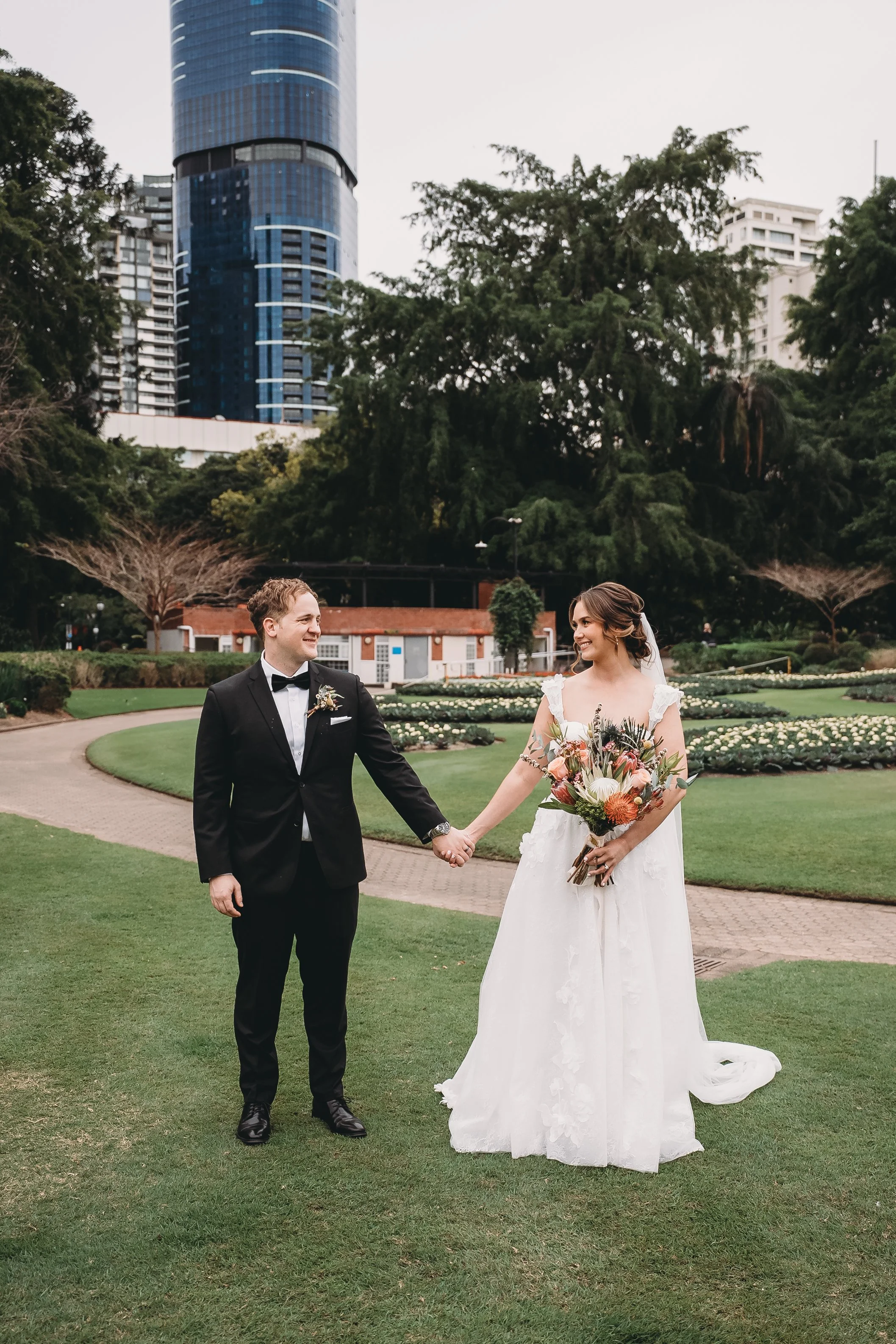A bride and groom holding hands in a park. The bride is in a white wedding dress holding a bouquet of flowers, and the groom is in a black tuxedo. Tall buildings are visible in the background.