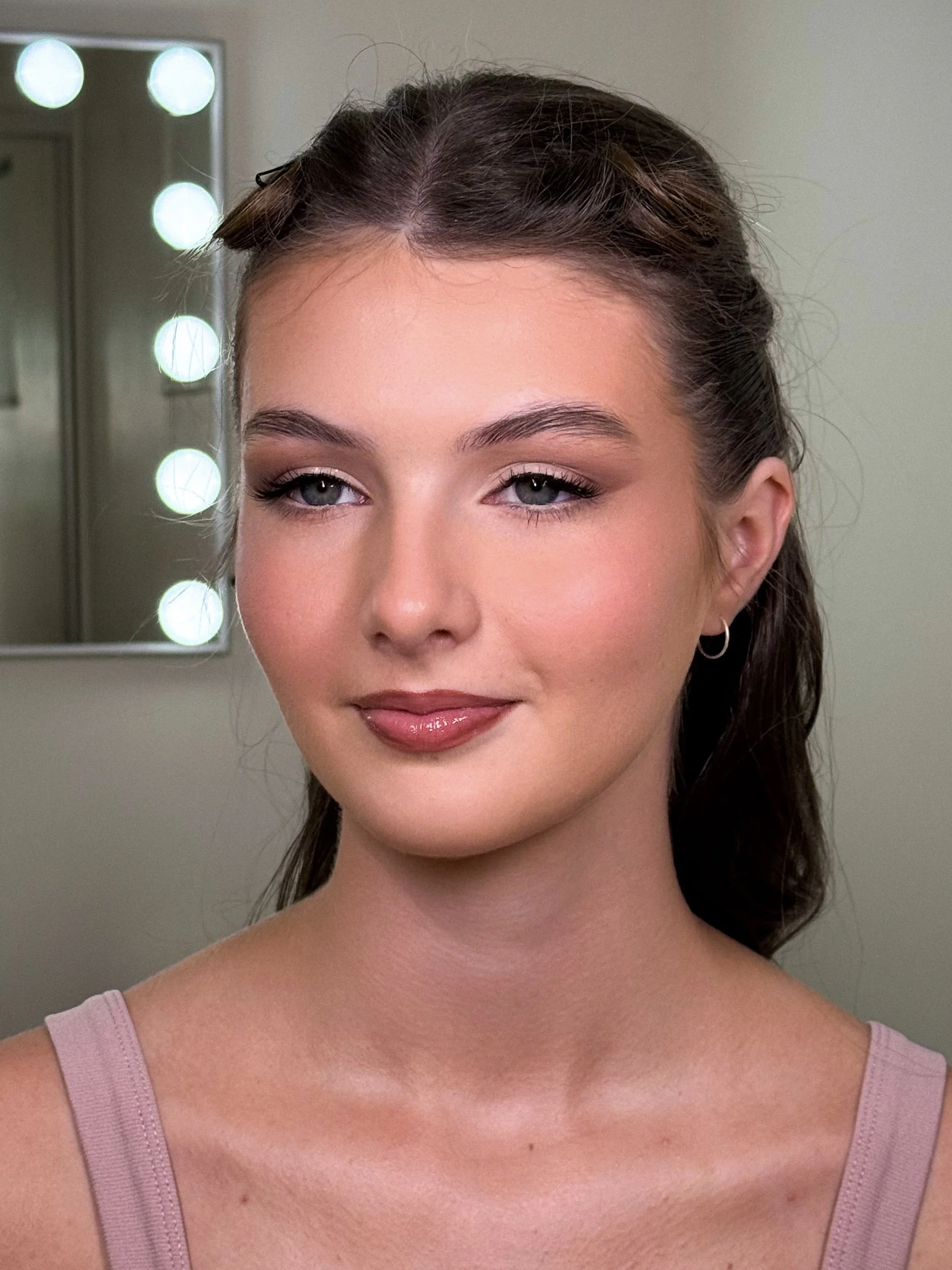 Close-up portrait of a young woman with light skin, blue eyes, and dark brown hair styled with soft waves. She is smiling subtly and wearing a pink sleeveless top and small hoop earrings, with a makeup look that includes natural tones, eyeliner, and 