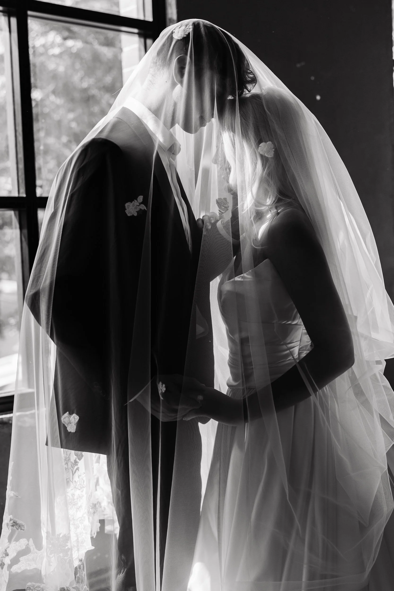 Black and white photo of a bride and groom standing close together, under a veil, holding hands, near a window with some natural light.