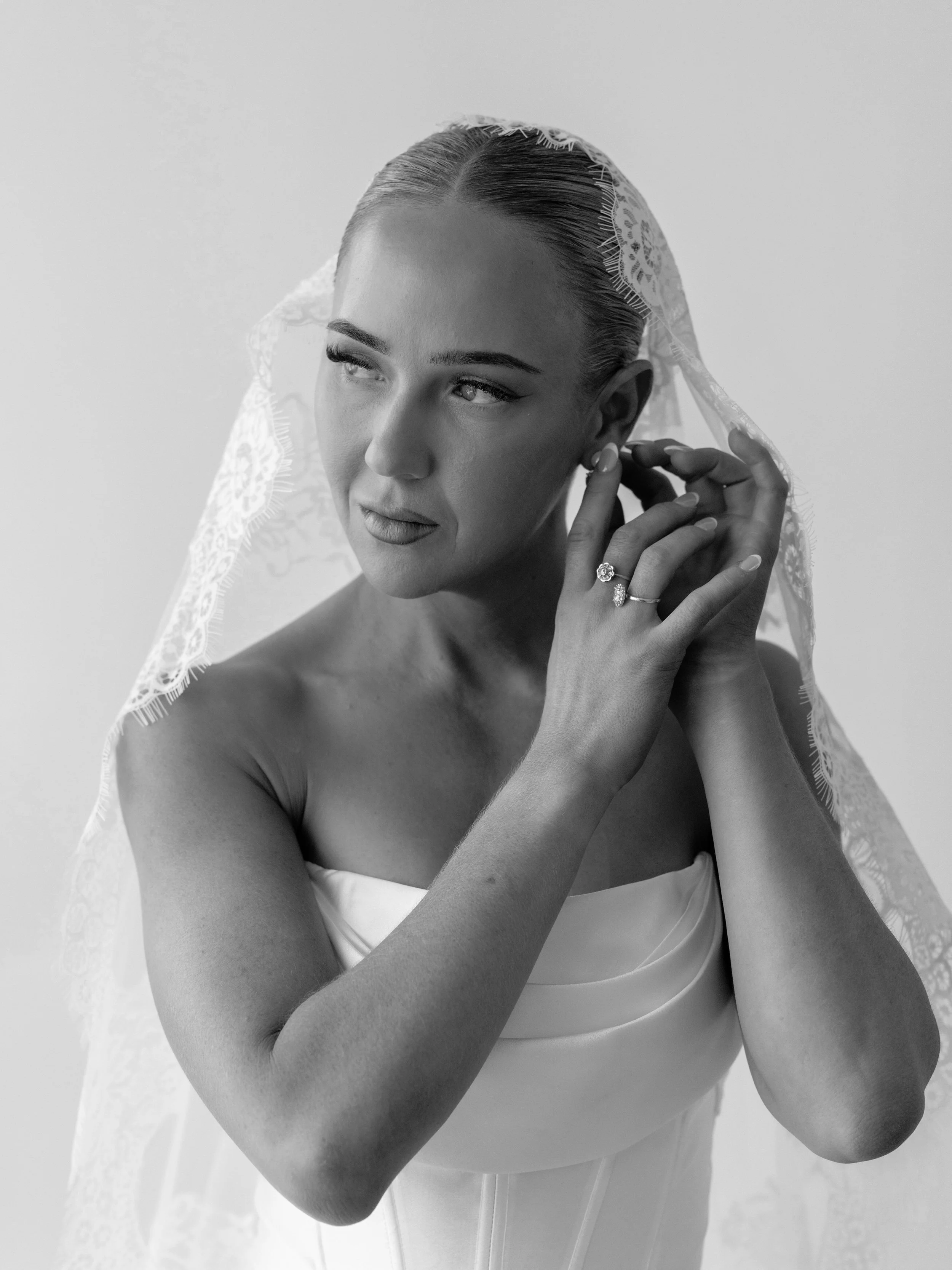 Black and white portrait of a woman in wedding attire, adjusting an earring while wearing a lace veil and rings.