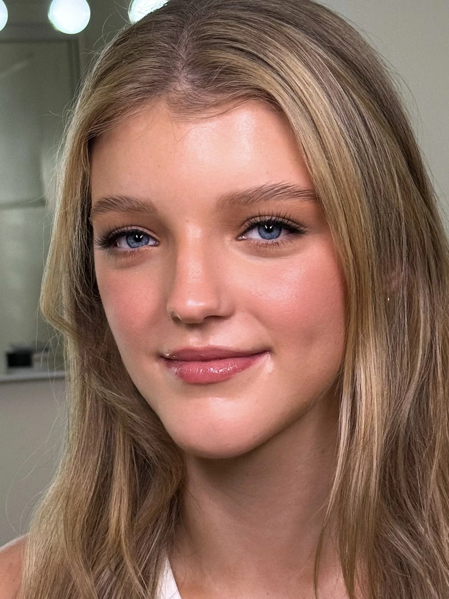 Close-up of a young woman with long, wavy blonde hair and blue eyes, smiling softly in a brightly lit room.