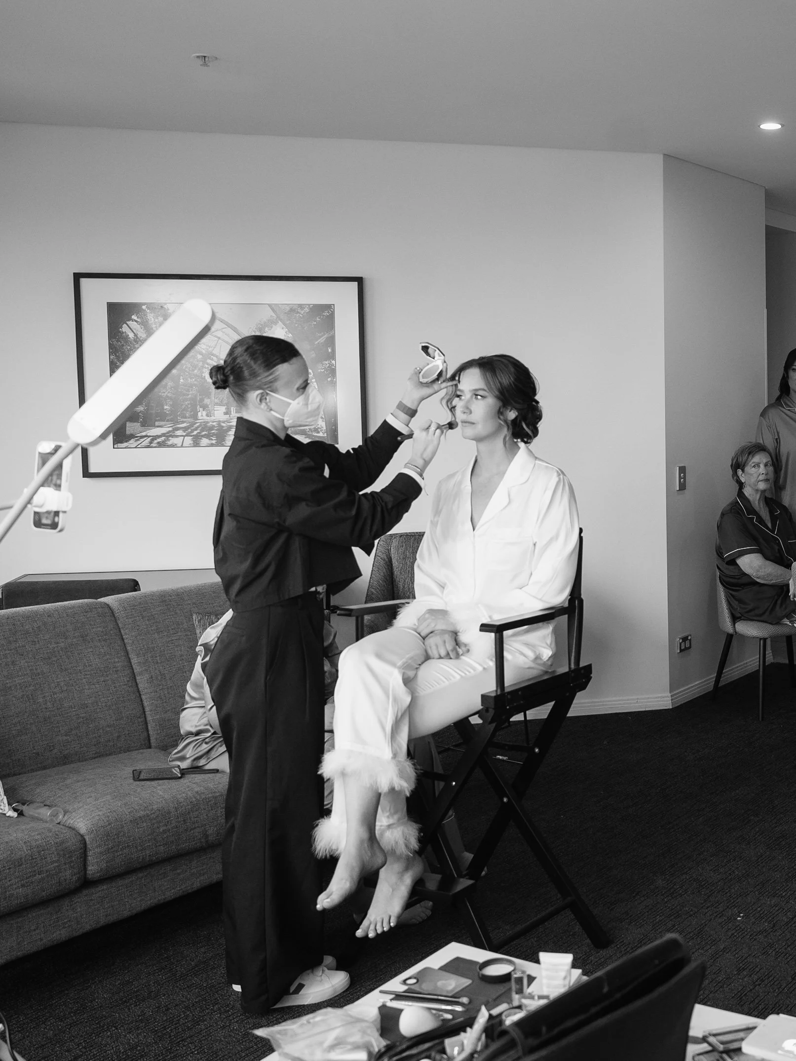 A woman sitting on a chair getting her makeup done by a makeup artist in a room, with a woman sitting on a couch in the background.