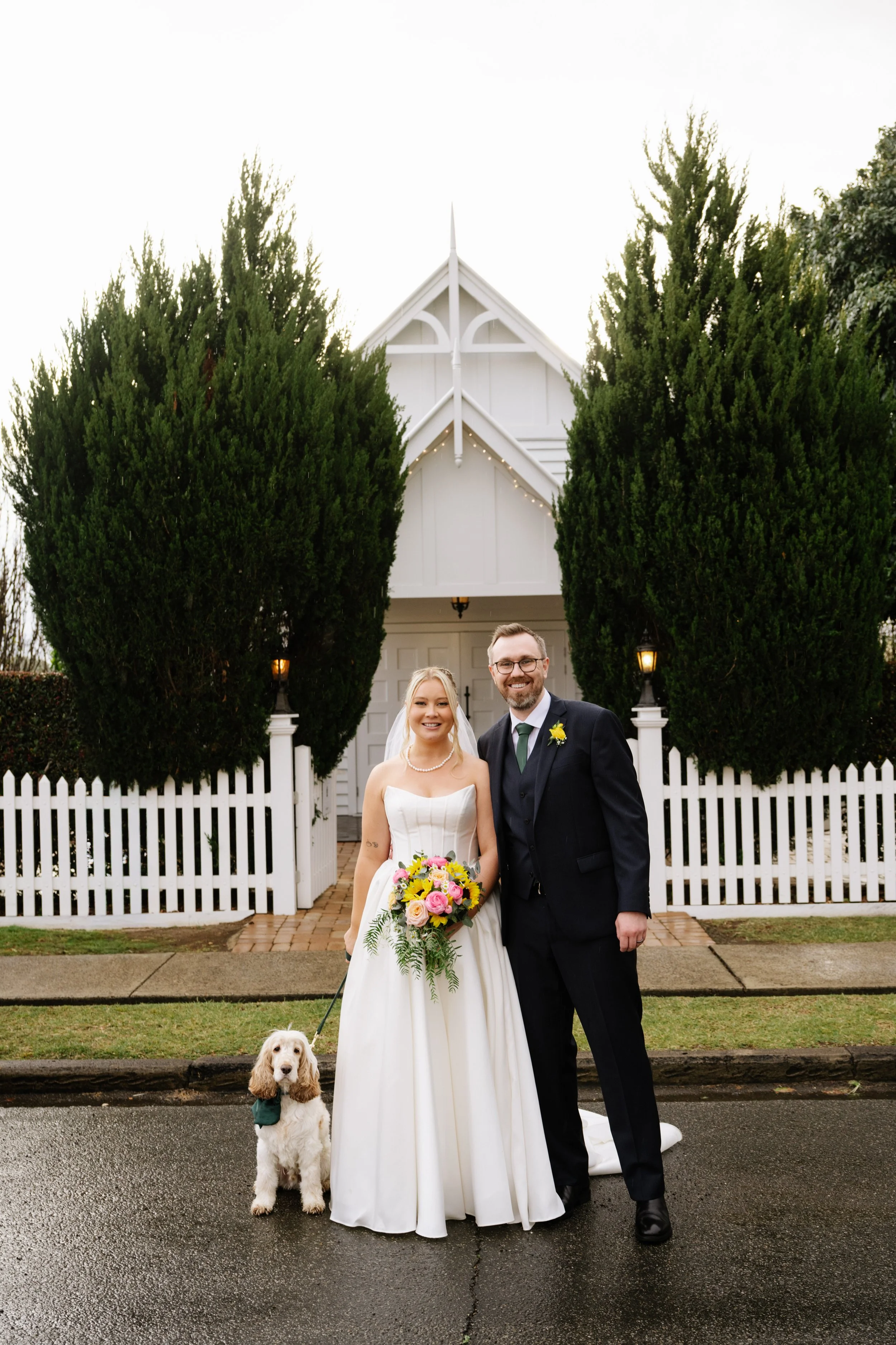 A bride and groom standing outside a white wedding chapel, smiling. The bride is holding a bouquet of flowers and is accompanied by a dog on a leash. The scene is on a wet street with a white picket fence and tall green trees in the background.