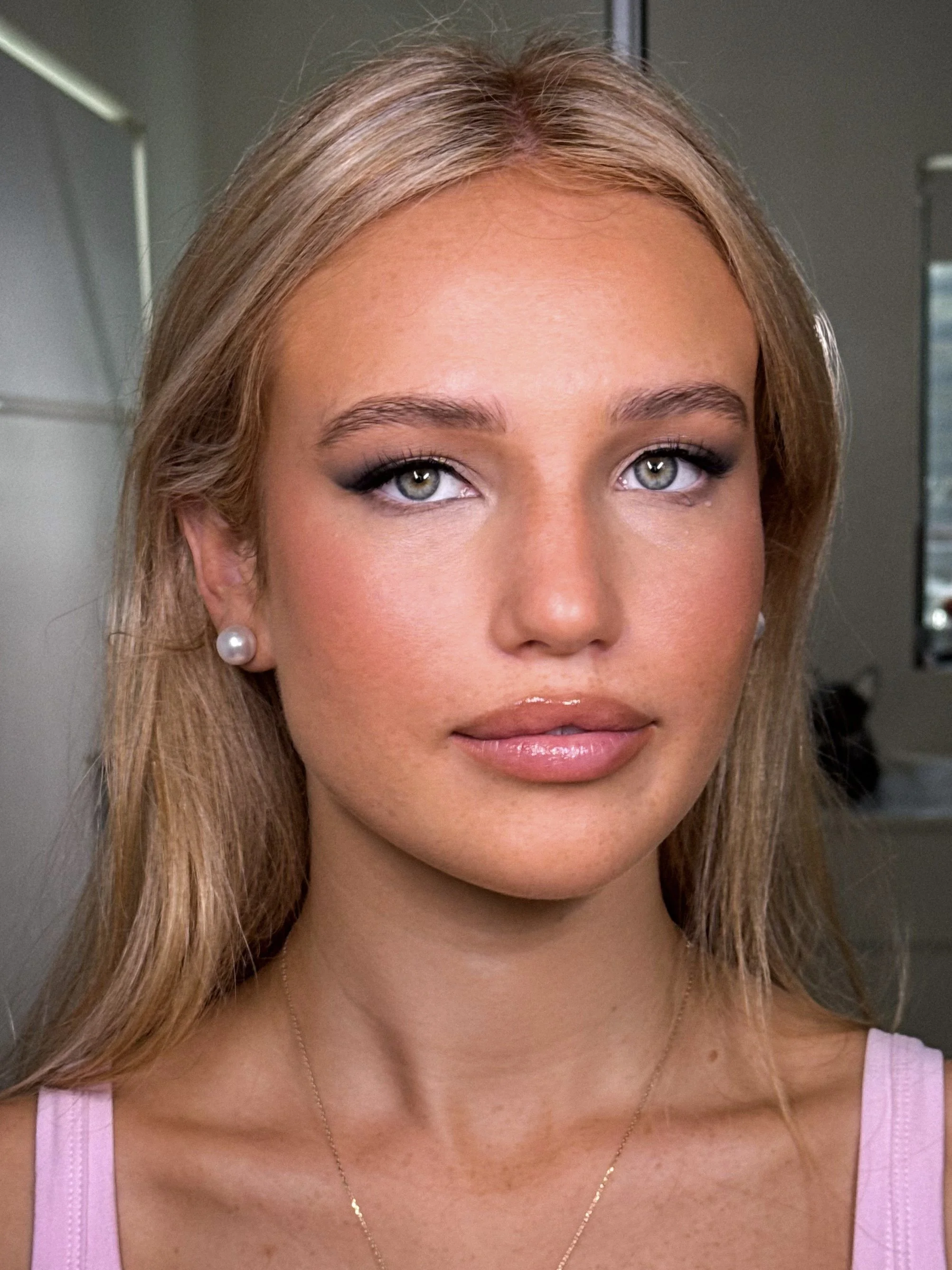 Close-up image of a young woman with blonde hair, blue eyes, wearing pearl earrings and a pink top, in an indoor setting.