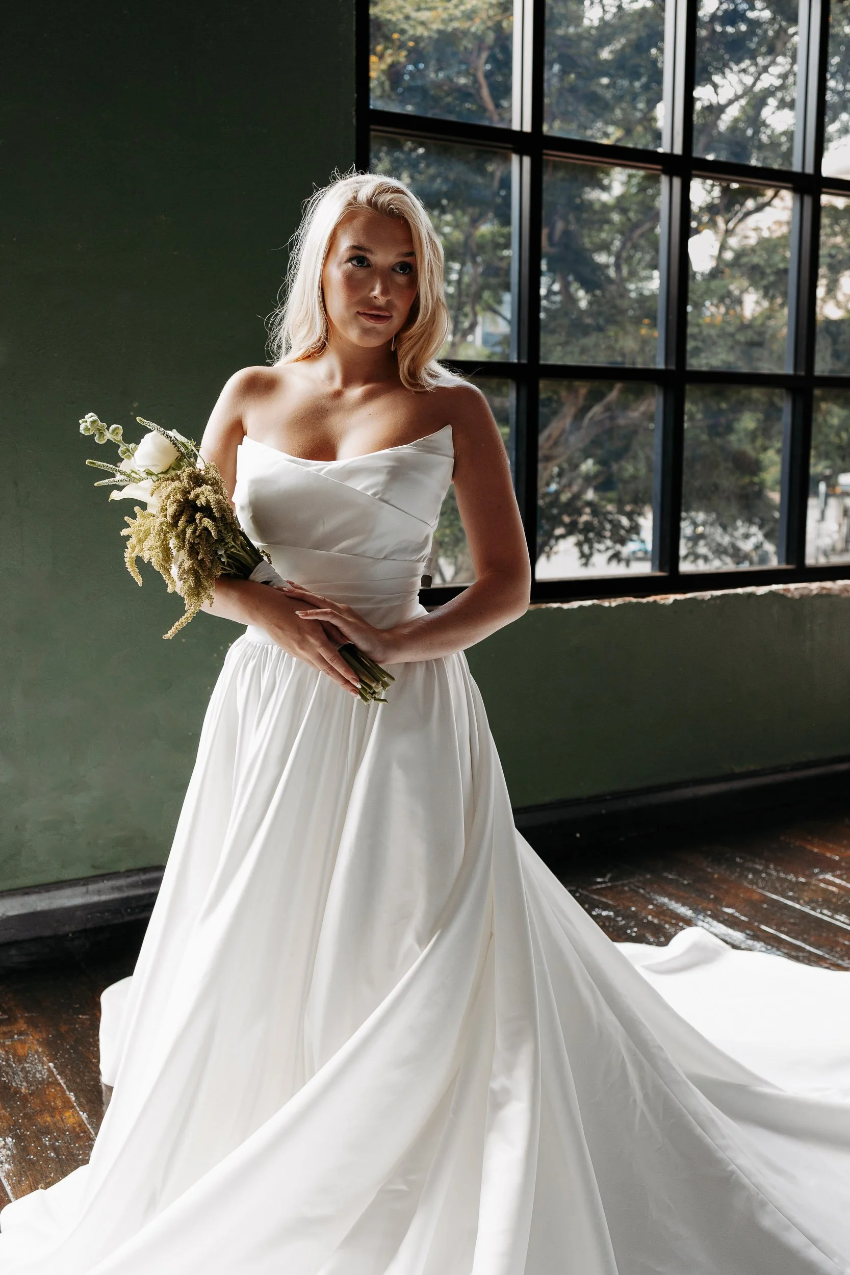 A bride in a white wedding dress holding a bouquet of flowers, standing indoors near a large window with trees outside.