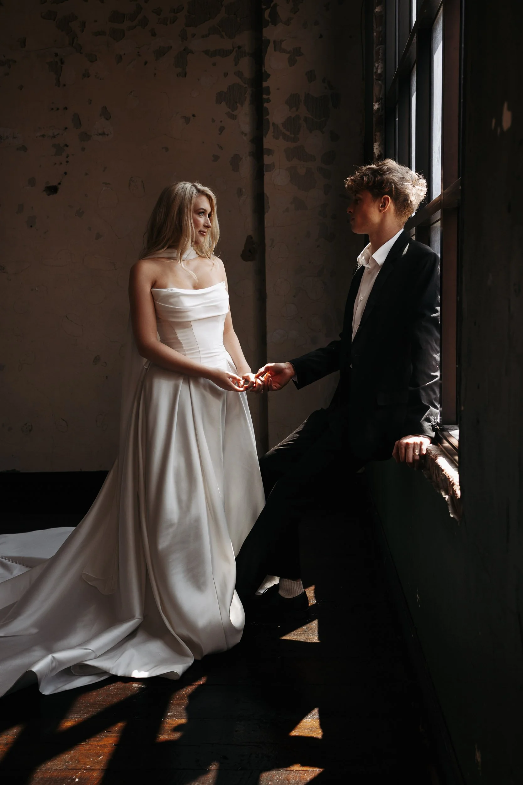 A bride and groom holding hands indoors, with sunlight streaming through a large window, the bride in a white wedding dress and the groom in a black suit.