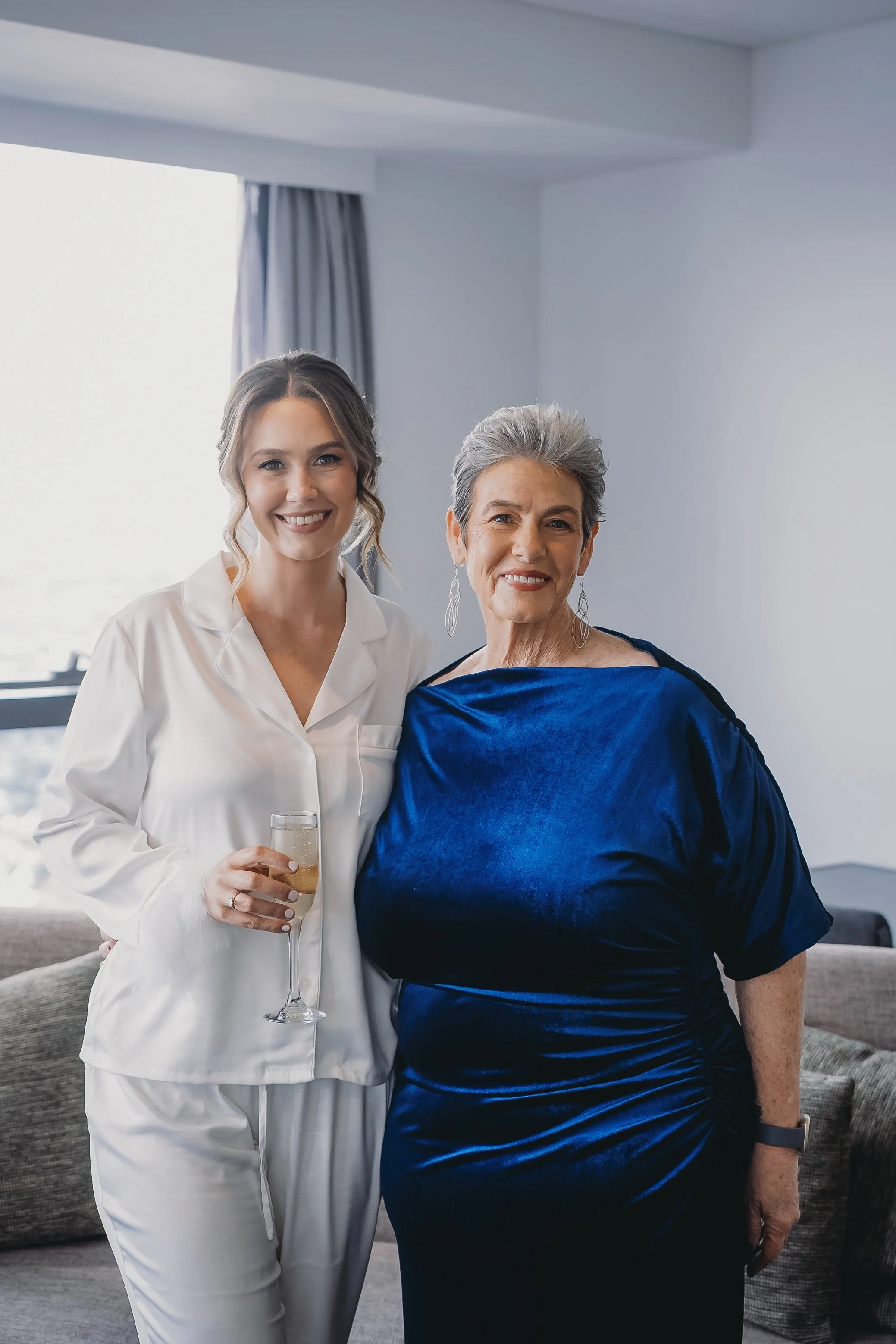 Two women smiling indoors, with a window and curtains in the background. The younger woman on the left is holding a glass of champagne and wearing white pajamas. The older woman on the right is wearing a shiny blue dress and earrings.