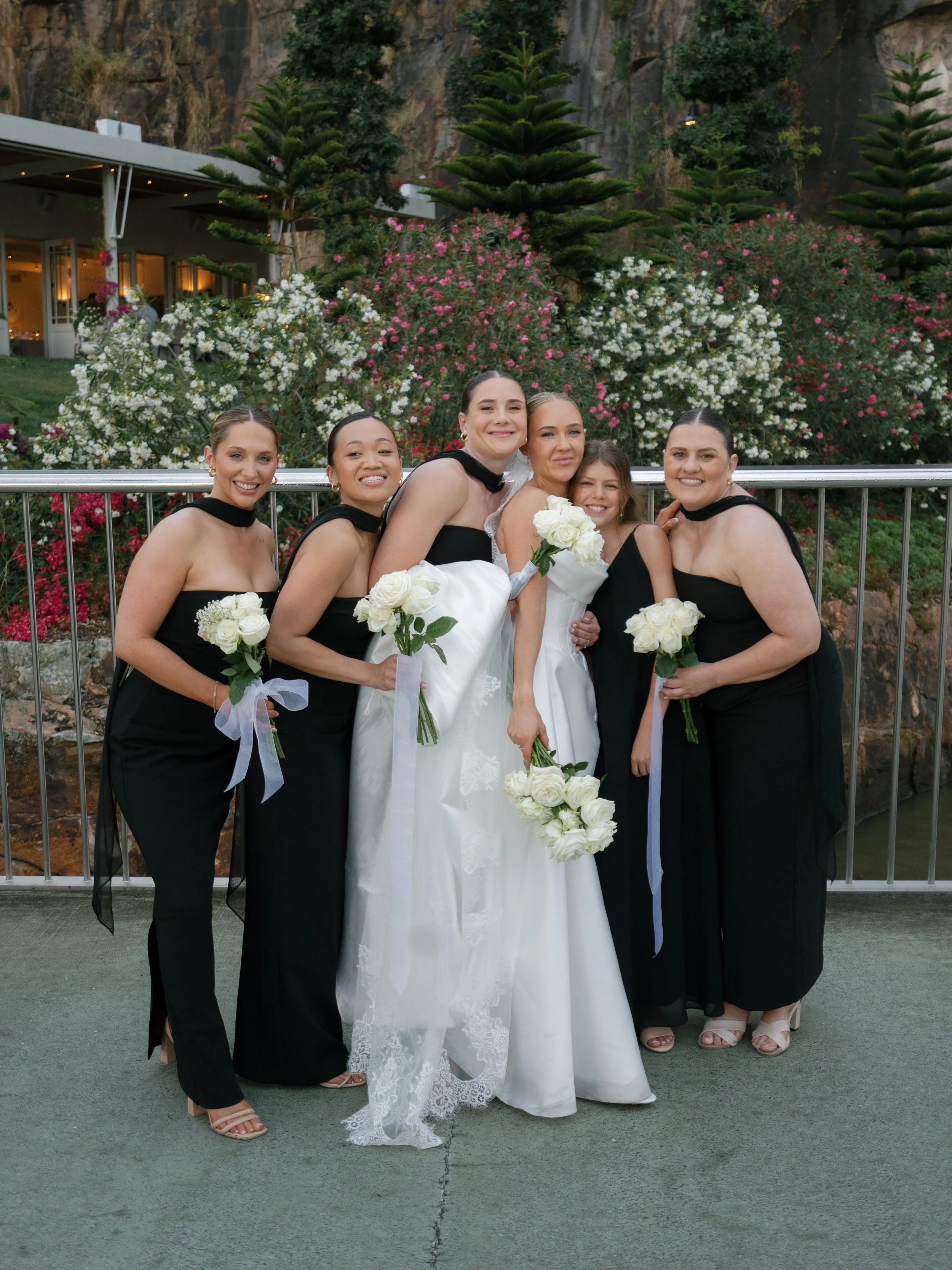 Group of six women, including a bride in a white wedding dress holding a bouquet, posing together outdoors with colorful flowers and a rocky hillside in the background.