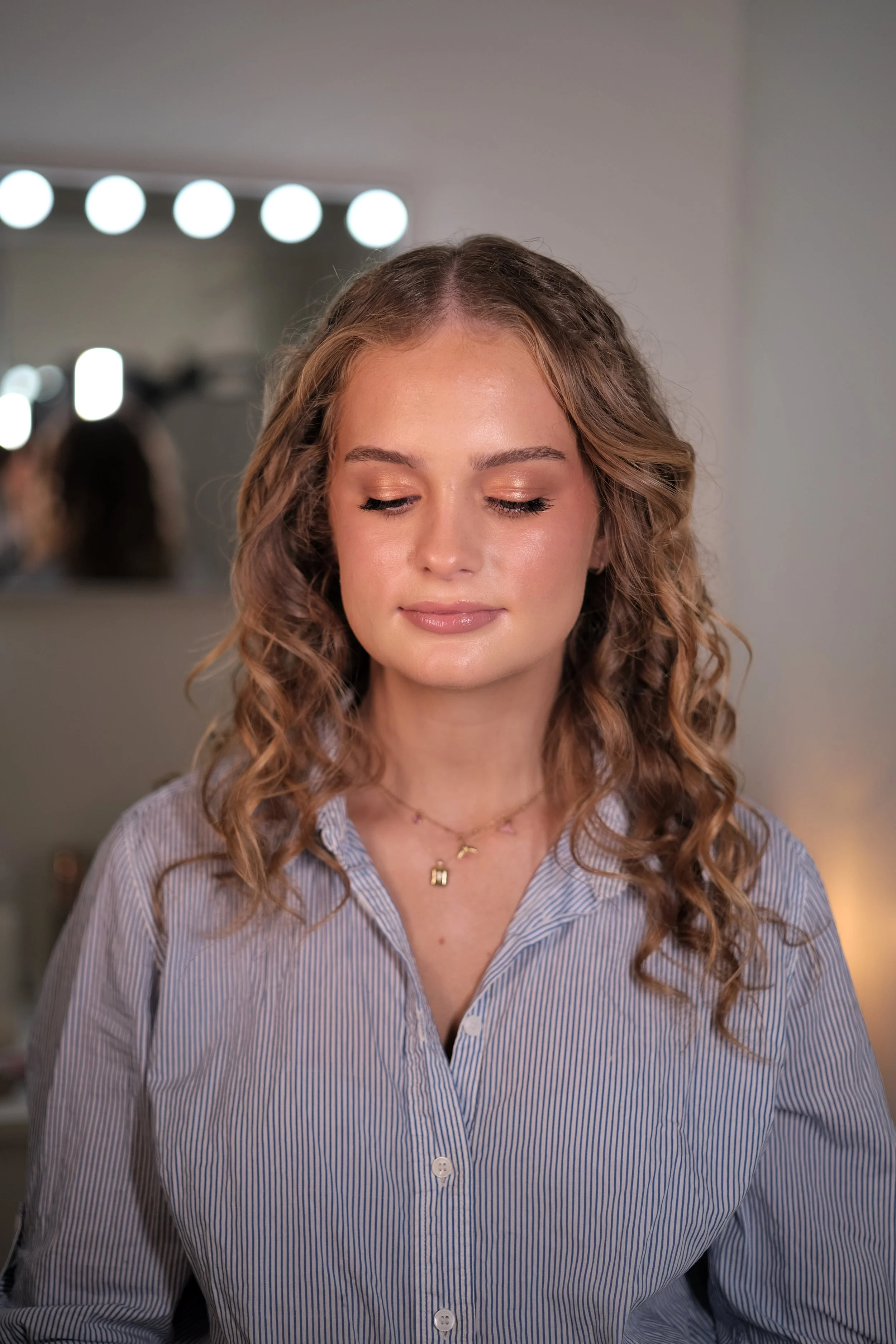 A woman with wavy, light brown hair and closed eyes, wearing a blue and white striped shirt and layered necklaces, standing in front of a mirror with bright lights.