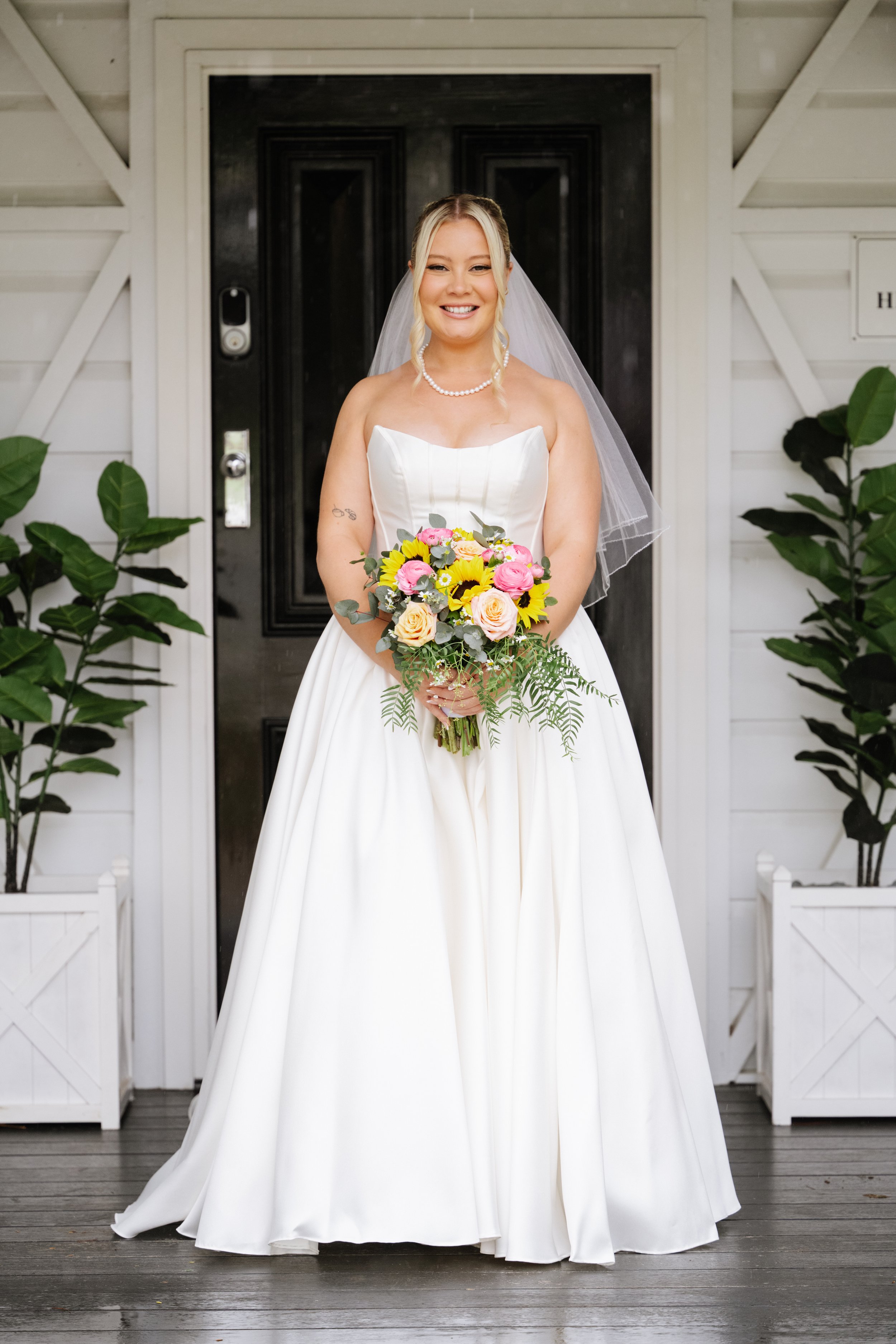 A smiling bride in a white wedding dress holding a colorful bouquet stands on a porch in front of a black door and white siding, flanked by potted plants.