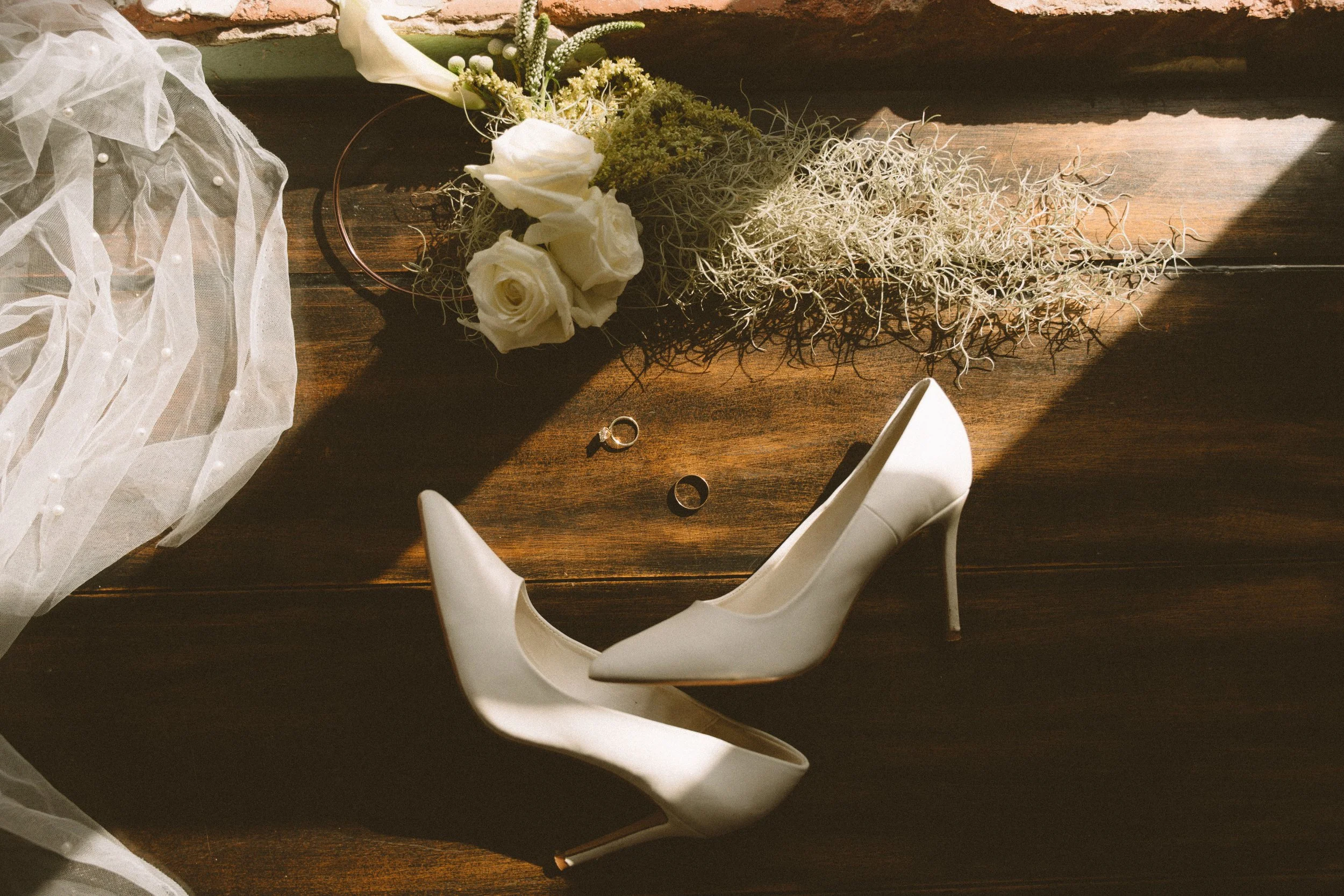 White high-heeled shoe, wedding rings, white roses, a bouquet of flowers, and sheer fabric with pearl details on a wooden surface with sunlight and shadow.