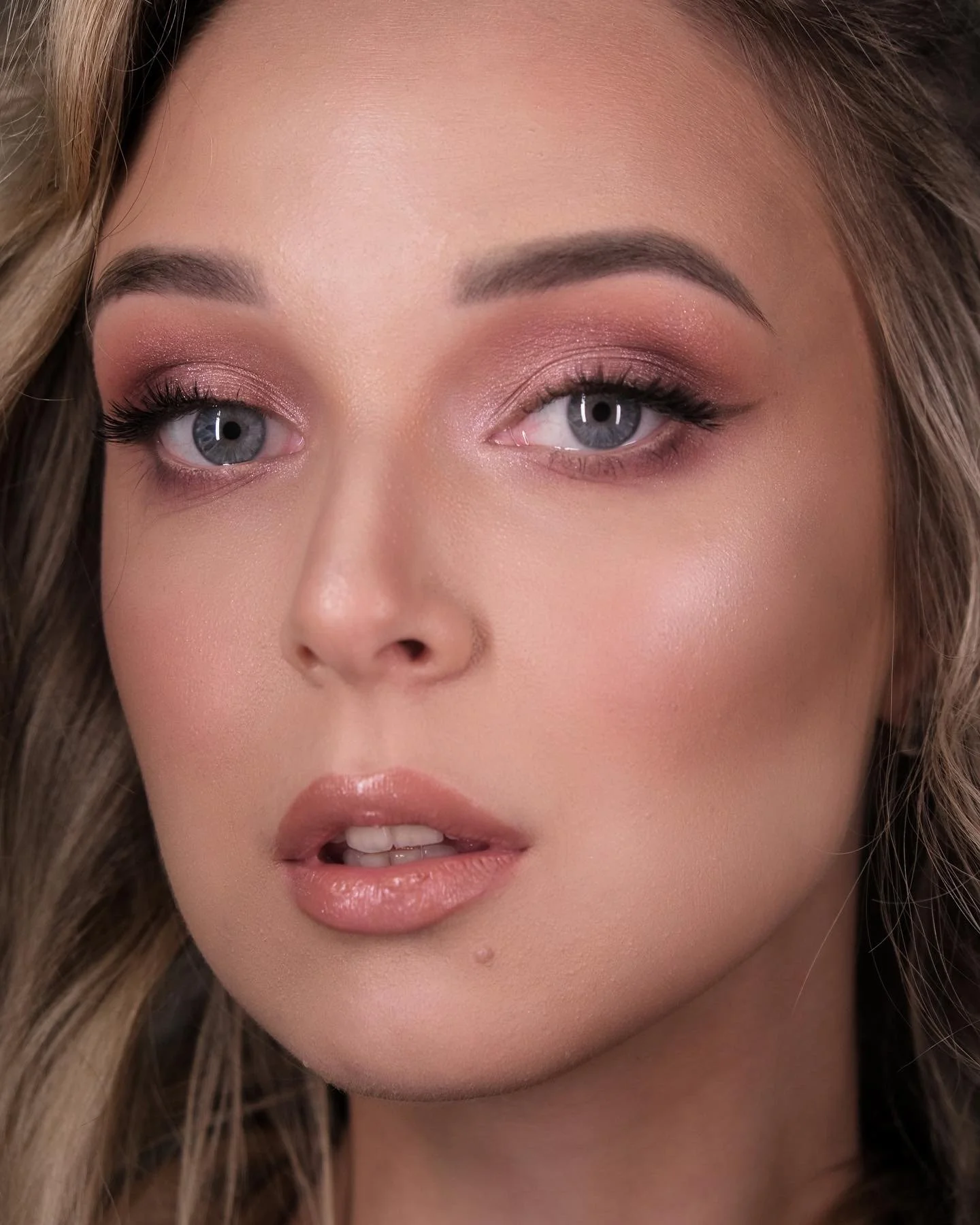 Close-up of a woman with blonde hair, light skin, blue eyes, and makeup including eyebrows, eyeshadow, mascara, blush, and glossy lips.