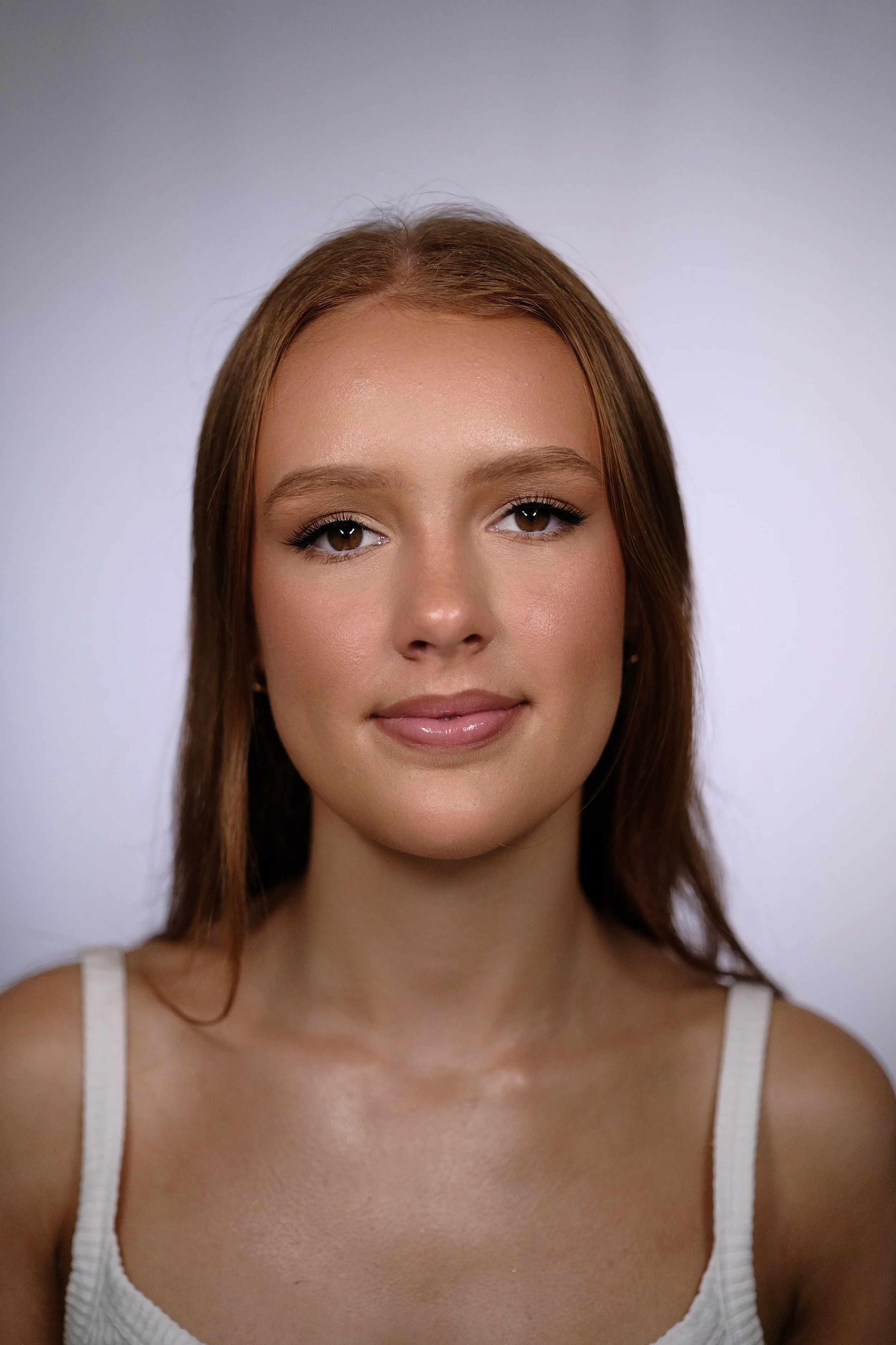 A close-up portrait of a young woman with long brown hair, wearing a white tank top, smiling softly against a plain light gray background.