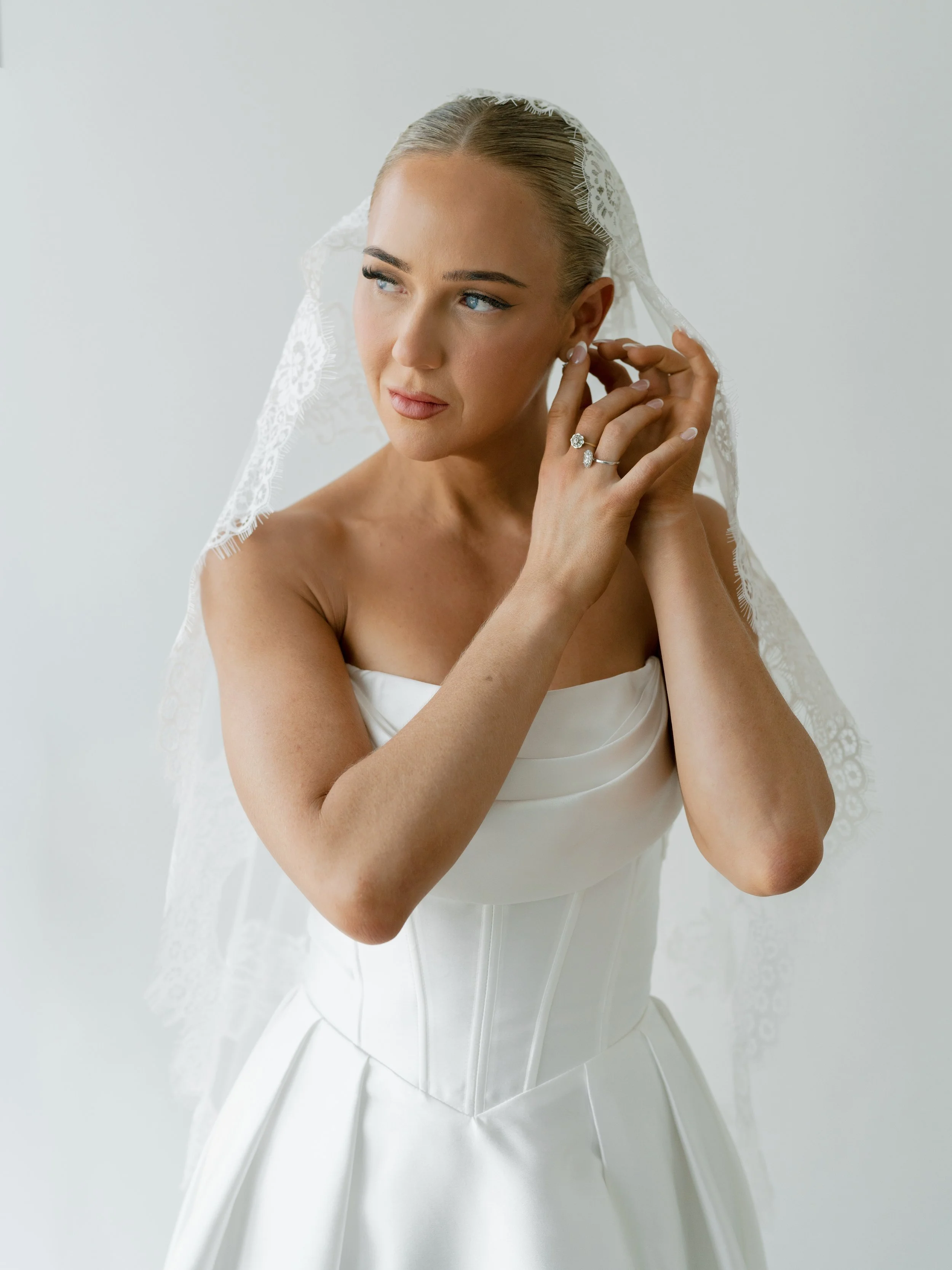 A bride touching her earring while wearing a white wedding dress and lace veil, with a ring on her finger, against a plain light background.