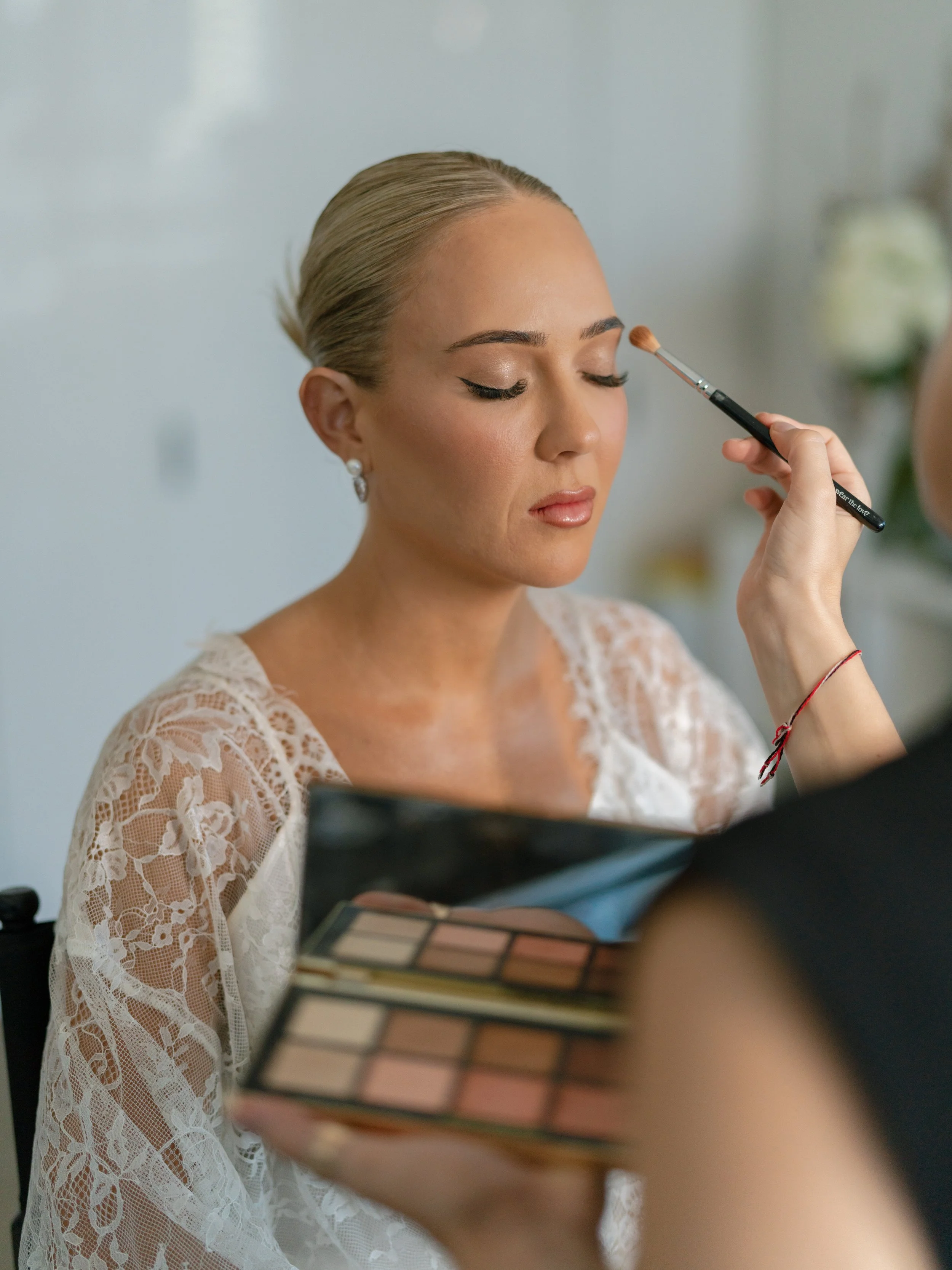 A woman in a lace dress has her makeup done, with a makeup artist applying eyeshadow to her eyelid and holding a makeup palette.