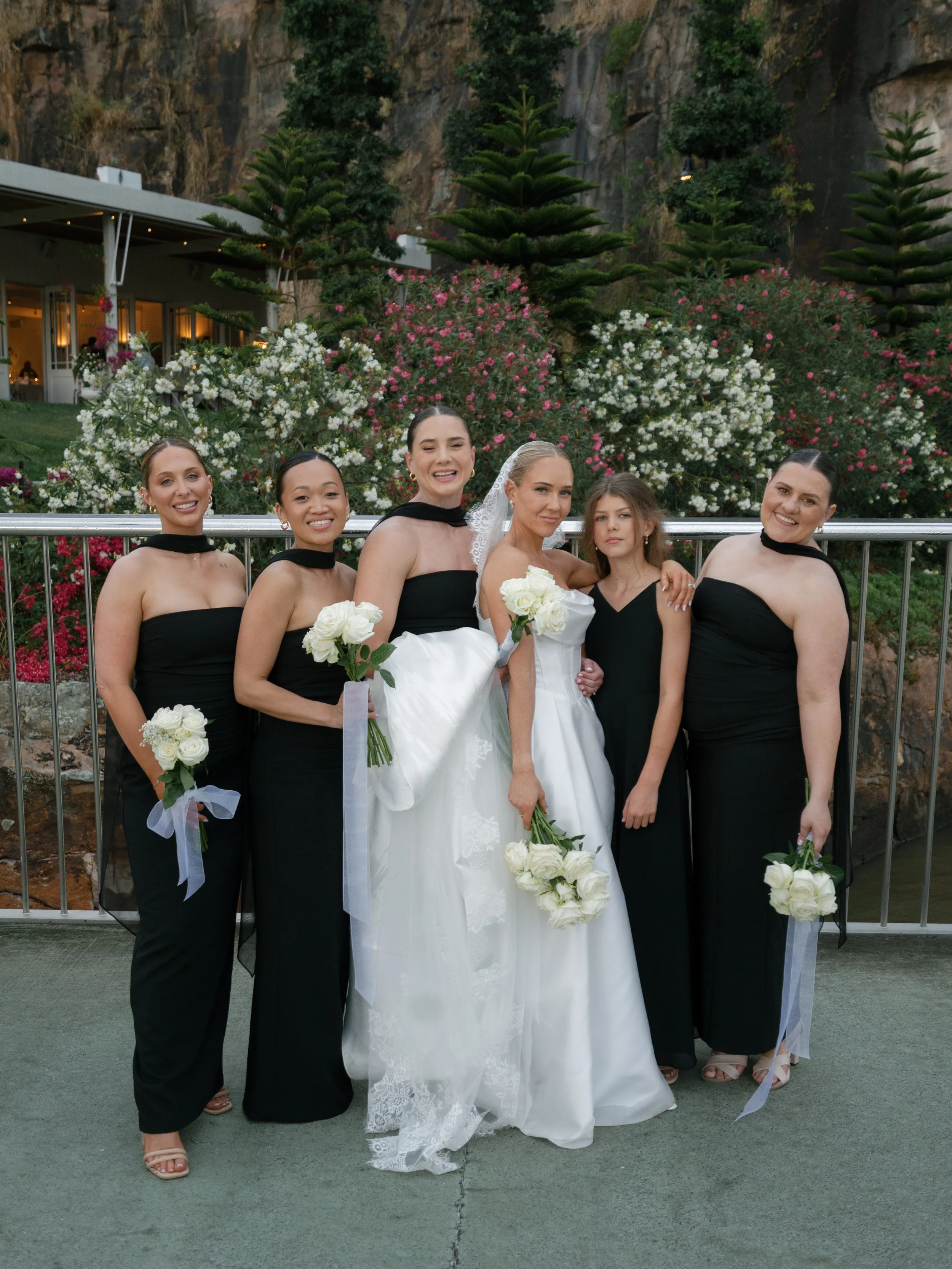 Group of six women, including a bride in a white gown, posing outdoors in front of flowering bushes and trees.