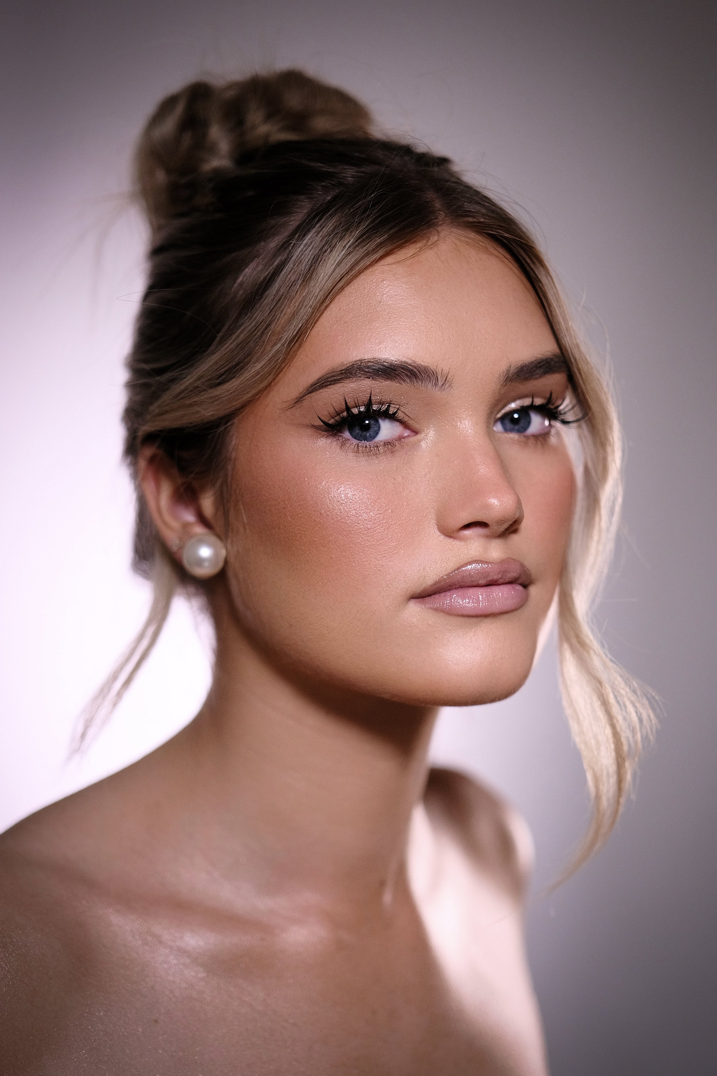 Close-up portrait of a young woman with blonde hair styled in a bun, wearing pearl earrings, and with makeup highlighting her blue eyes and glossy lips, against a neutral background.