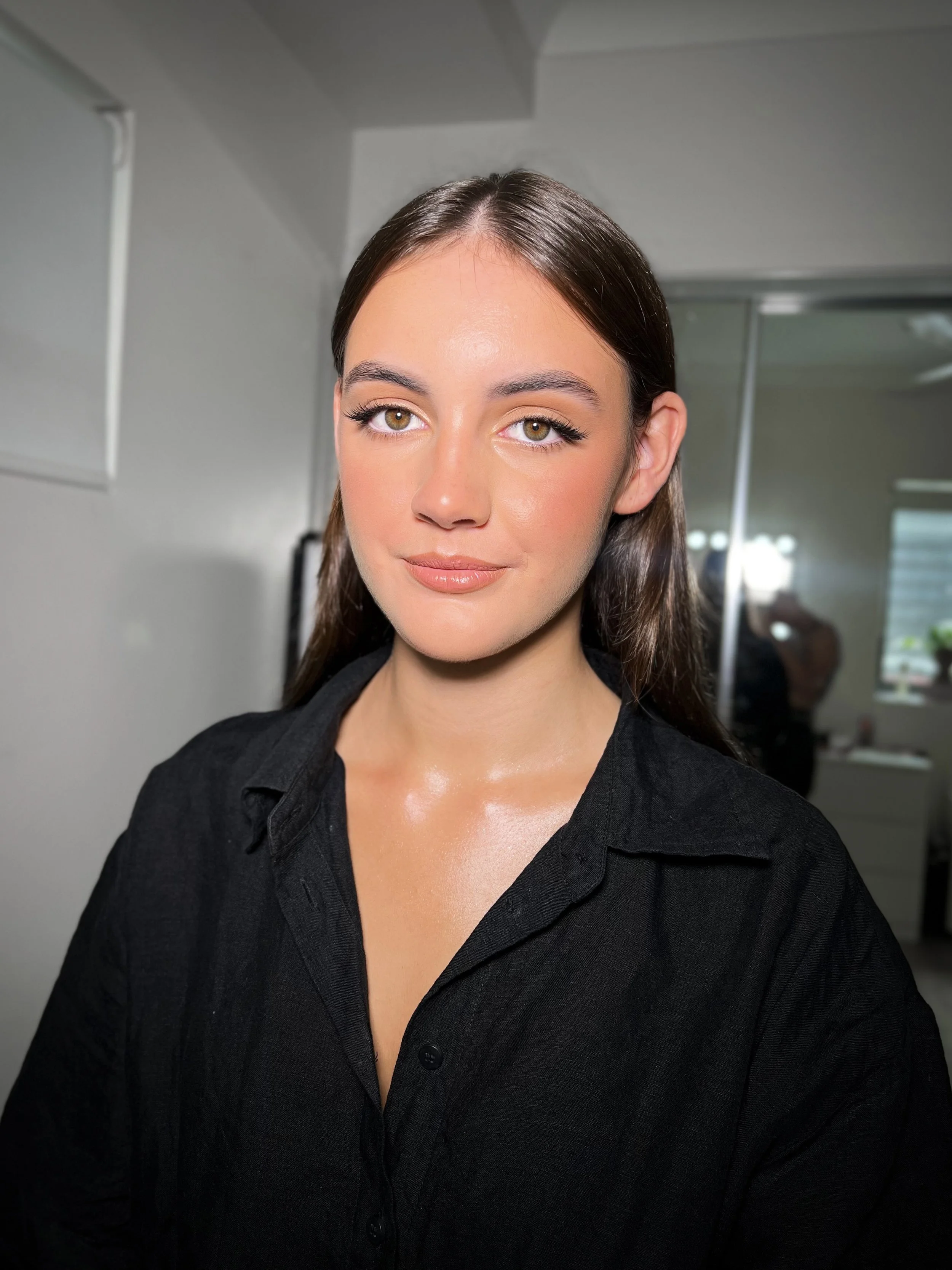 A young woman with long brown hair, wearing a black shirt, stands indoors with a plain background and a mirror behind her.