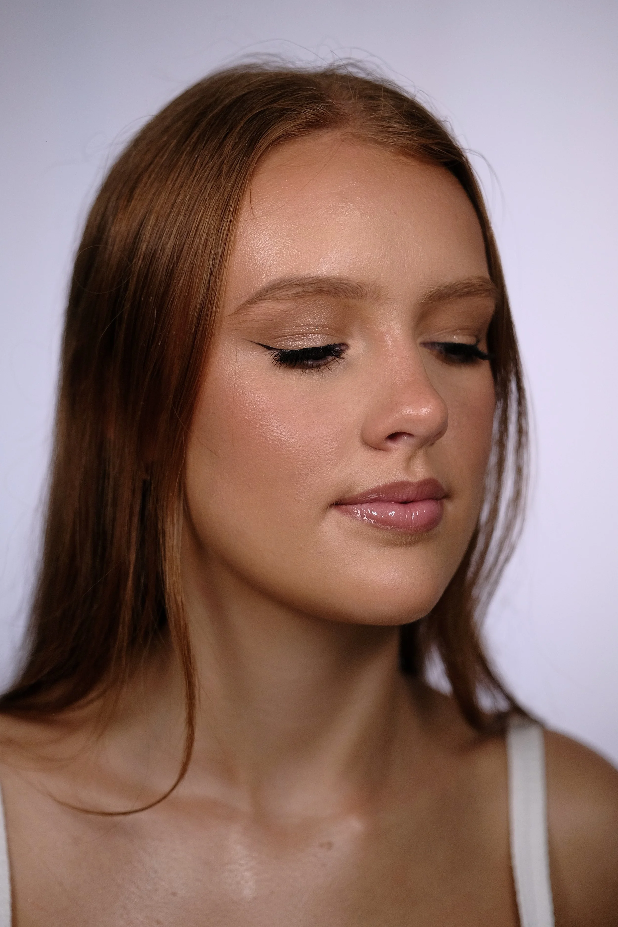 Close-up of a young woman with long auburn hair, wearing natural makeup with winged eyeliner, light eyeshadow, and glossy lips, against a plain light background.