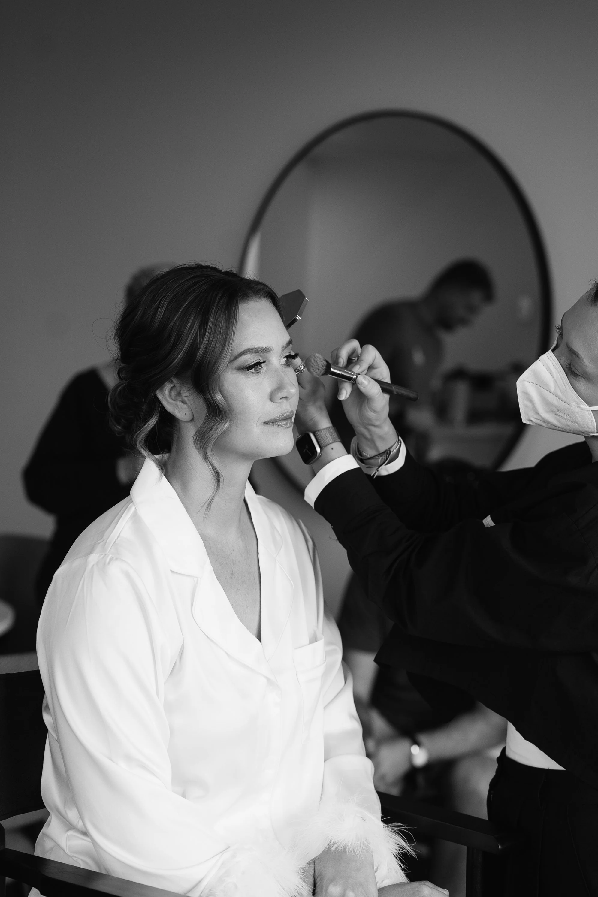 A woman sitting on a chair getting her makeup applied by a makeup artist. The woman is wearing a white shirt and the makeup artist is wearing a face mask.