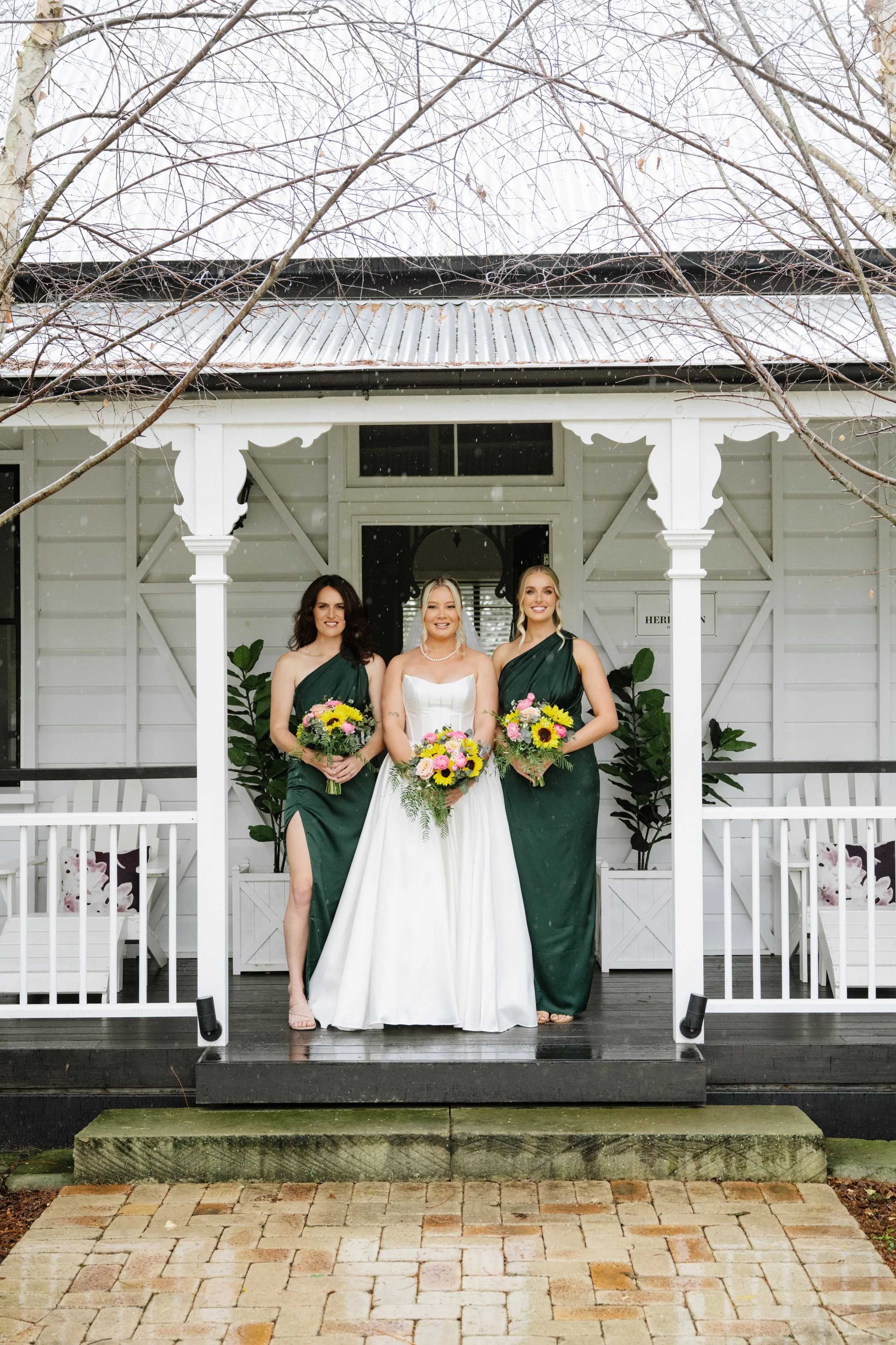 A bride in a white wedding gown and two bridesmaids in dark green dresses stand on a porch holding bouquets of colorful flowers.