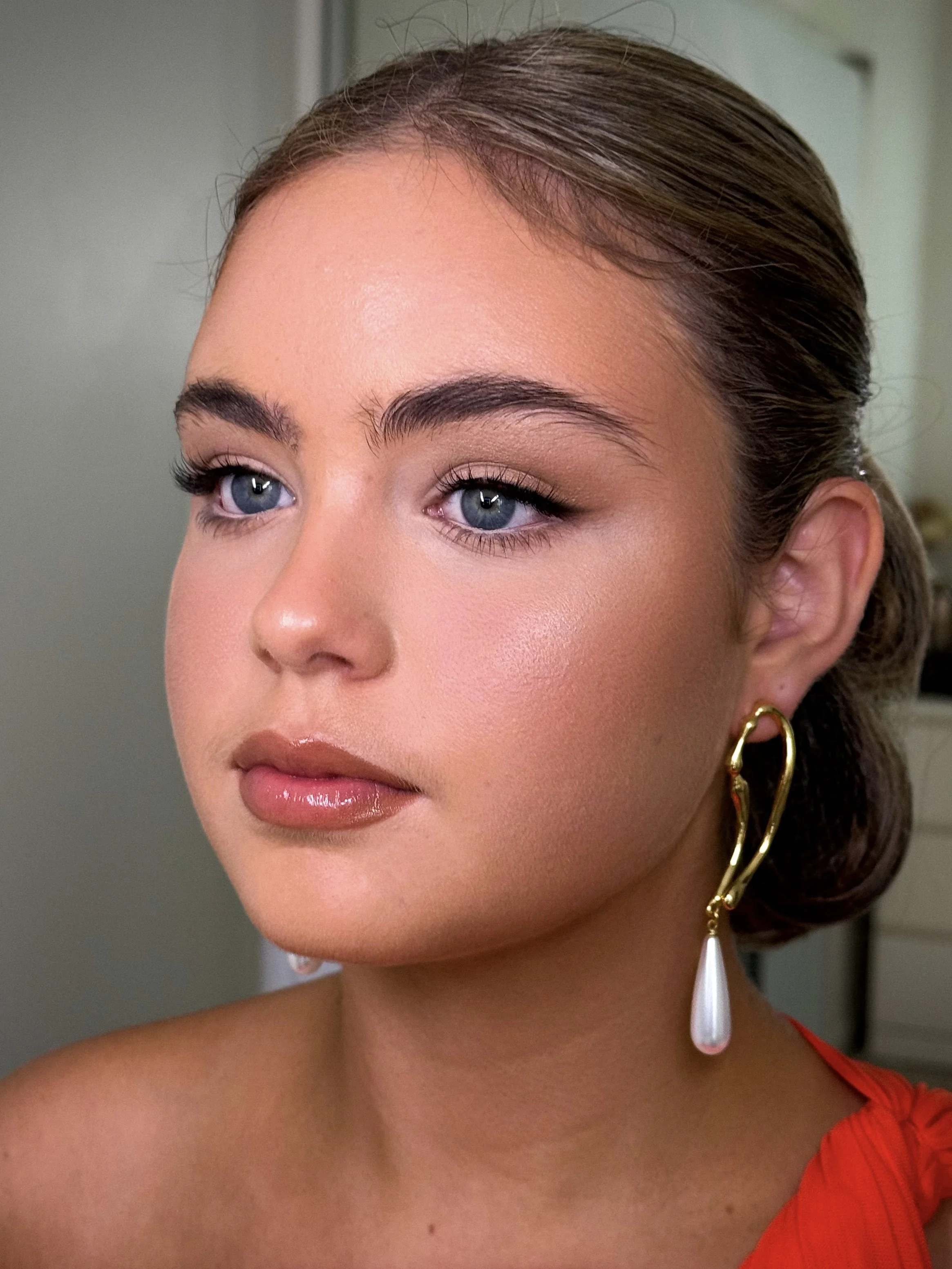 Close-up of a woman with blue eyes, brown hair styled in an elegant updo, wearing a red dress and a large gold earring with a pearl drop.