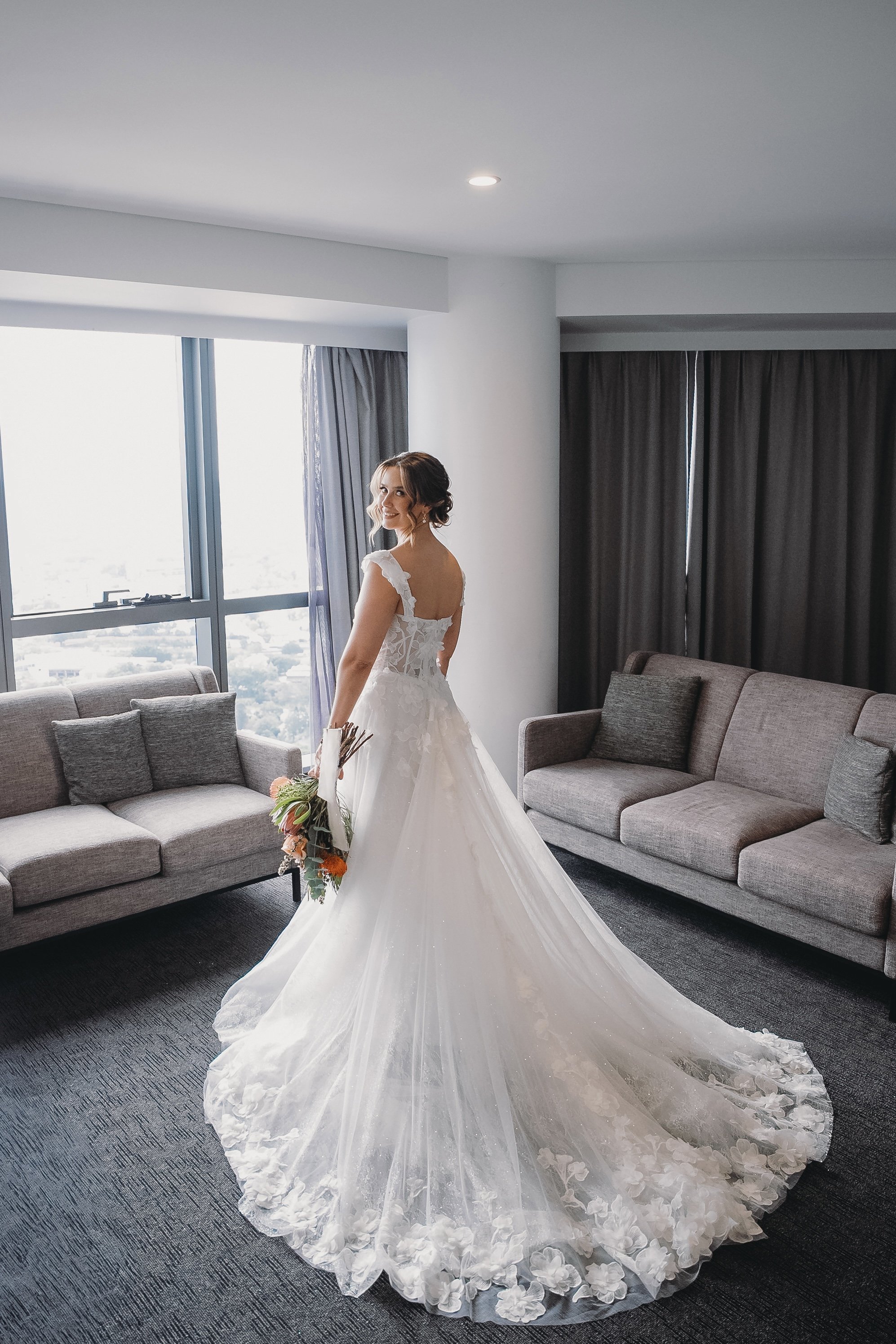 A bride in a wedding dress standing in a modern hotel room near a window, holding a bouquet of flowers, with gray sofas and dark curtains in the background.