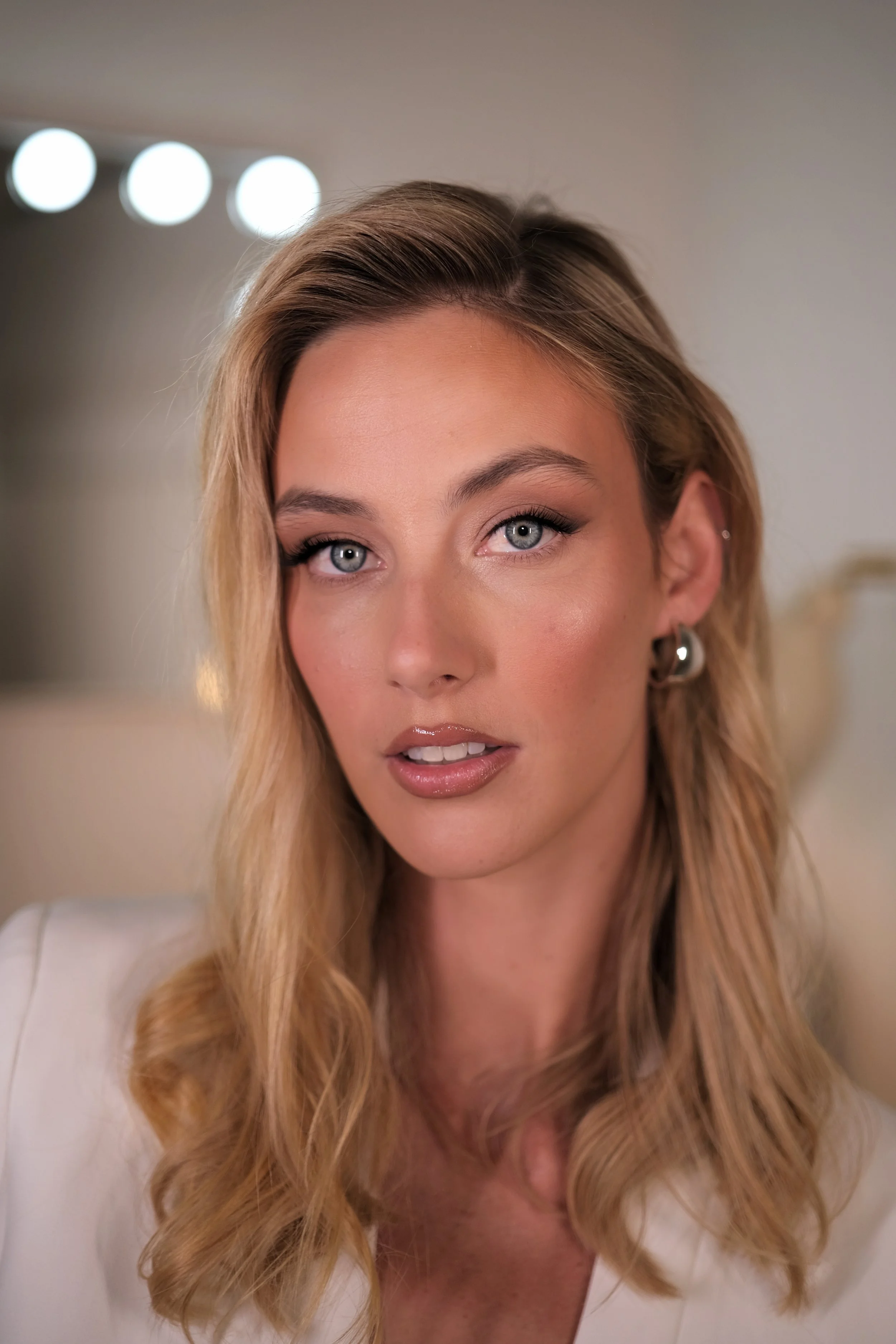 A woman with long blonde hair and blue eyes looking directly at the camera, wearing makeup and earrings, indoors with blurred lights in the background.
