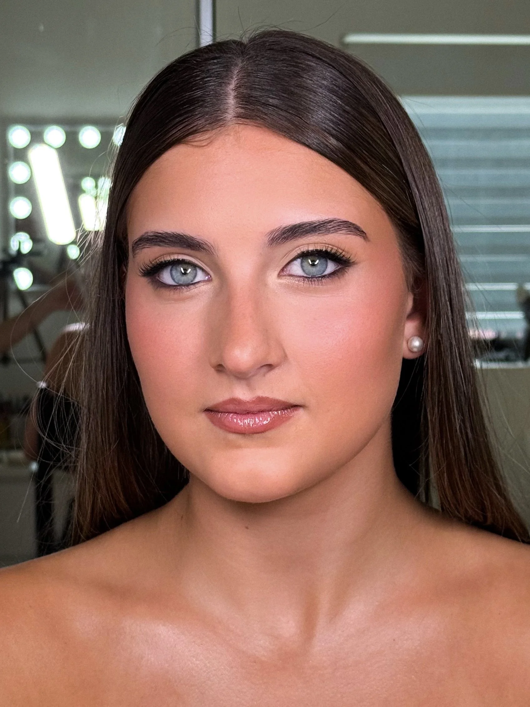Close-up of a young woman with long brown hair, blue eyes, pearl earrings, and subtle makeup, in a studio setting.