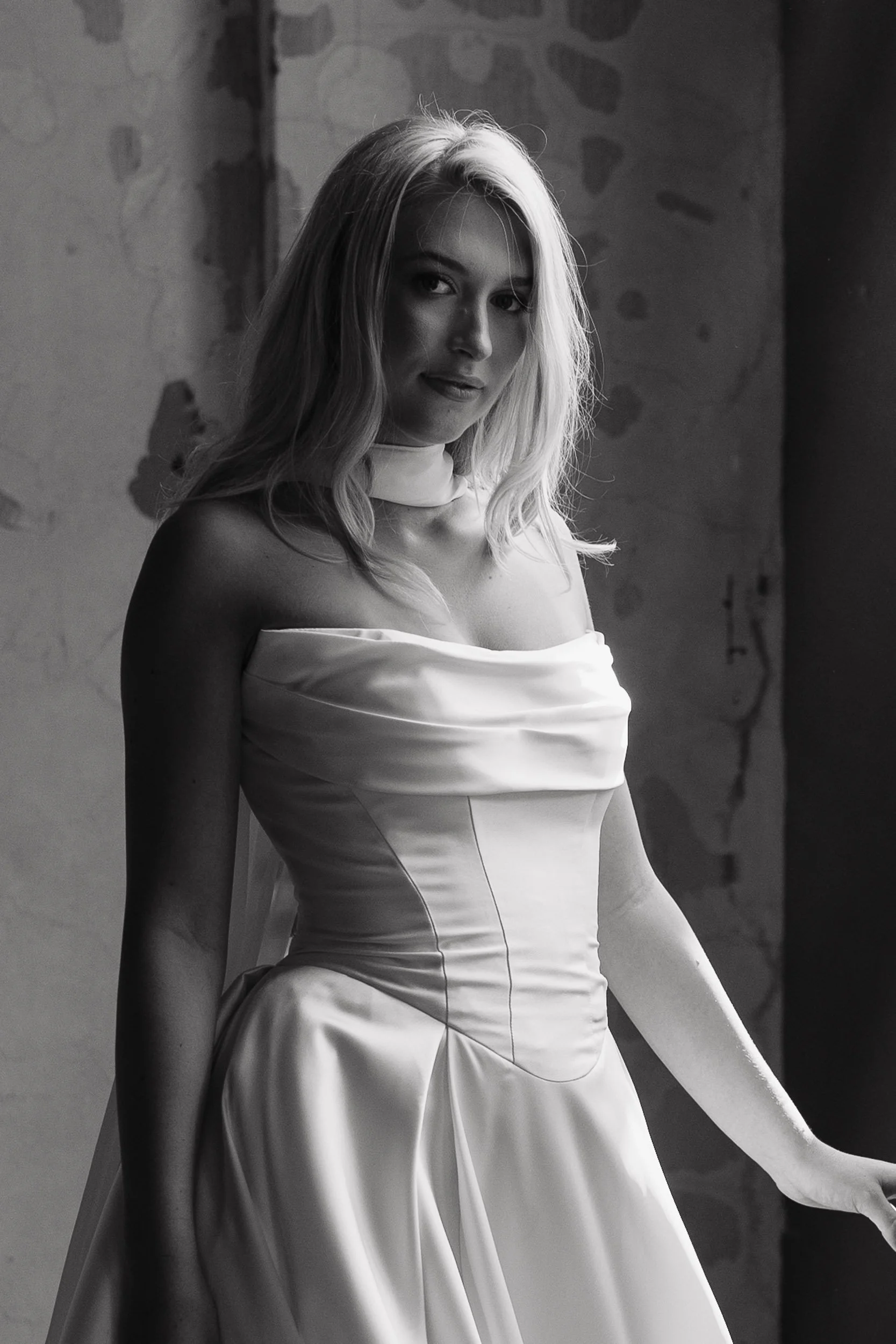 A woman in an off-shoulder satin dress standing in front of a textured wall, looking at the camera with a slight smile.