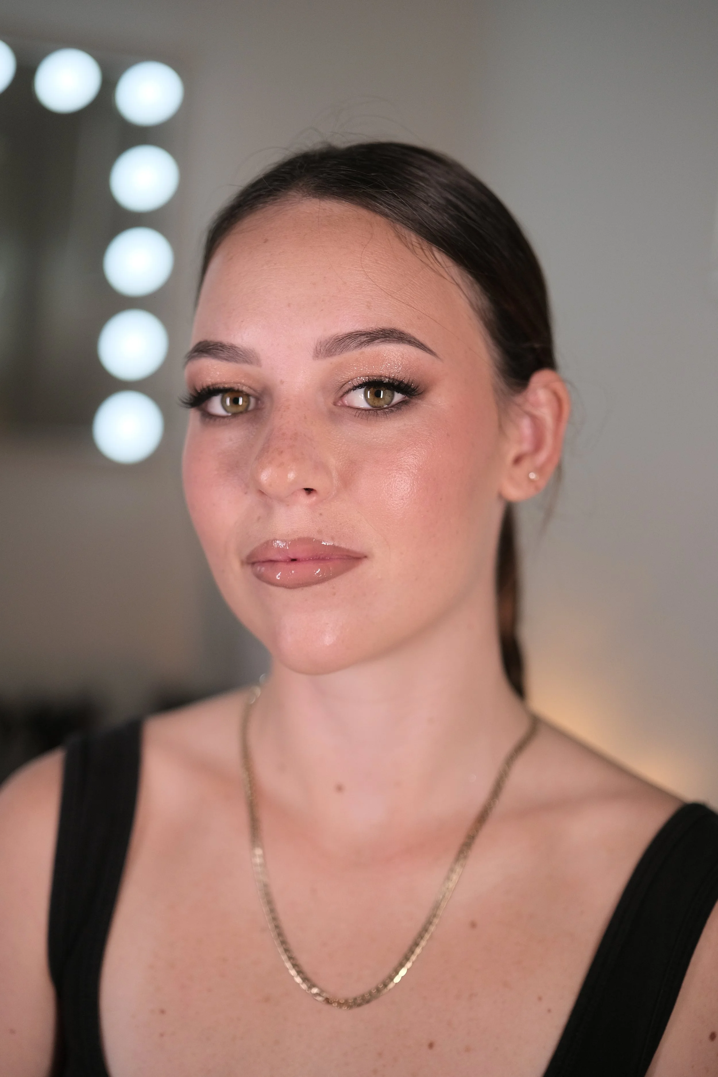 Close-up of a woman with brown hair, wearing makeup, a black top, and a gold necklace, in a room with a mirror and light bulbs.