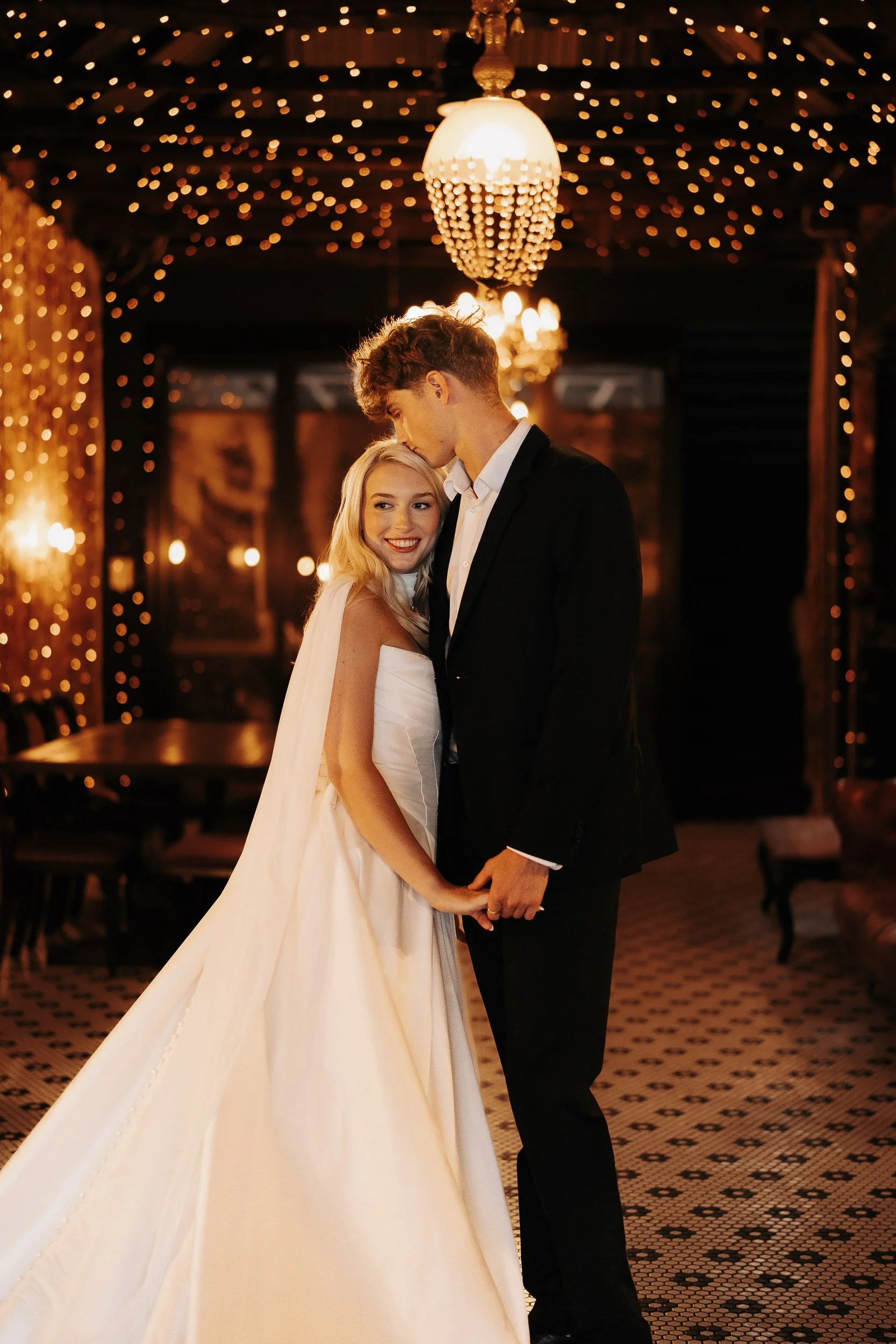 A bride and groom holding hands and embracing in a warmly lit, decorated venue with string lights and chandeliers at their wedding reception.