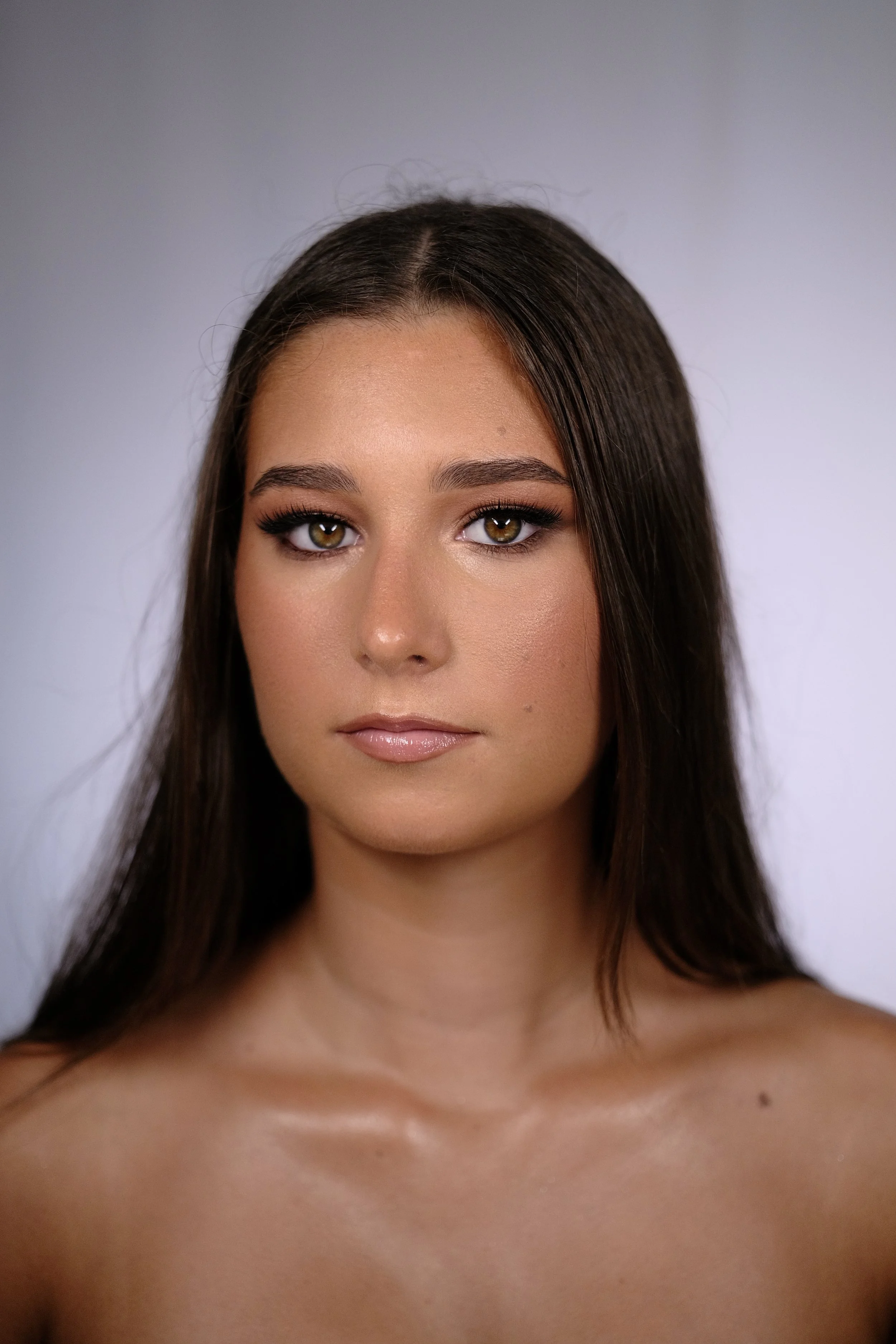 A portrait of a young woman with long brown hair, hazel eyes, and natural makeup, looking directly at the camera against a plain gray background.