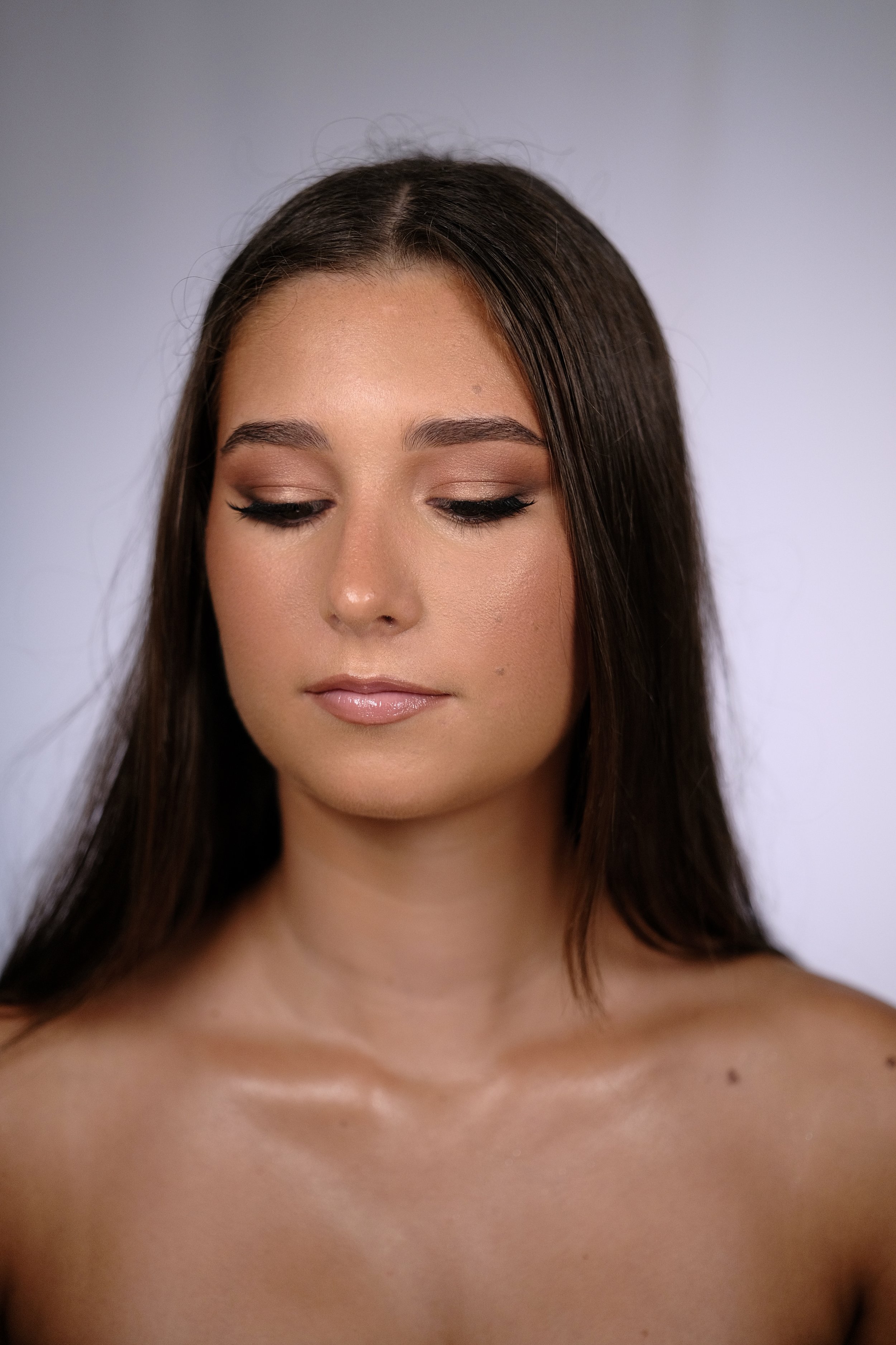 Close-up of a young woman with long brown hair, closed eyes, natural makeup, and bare shoulders against a neutral background.