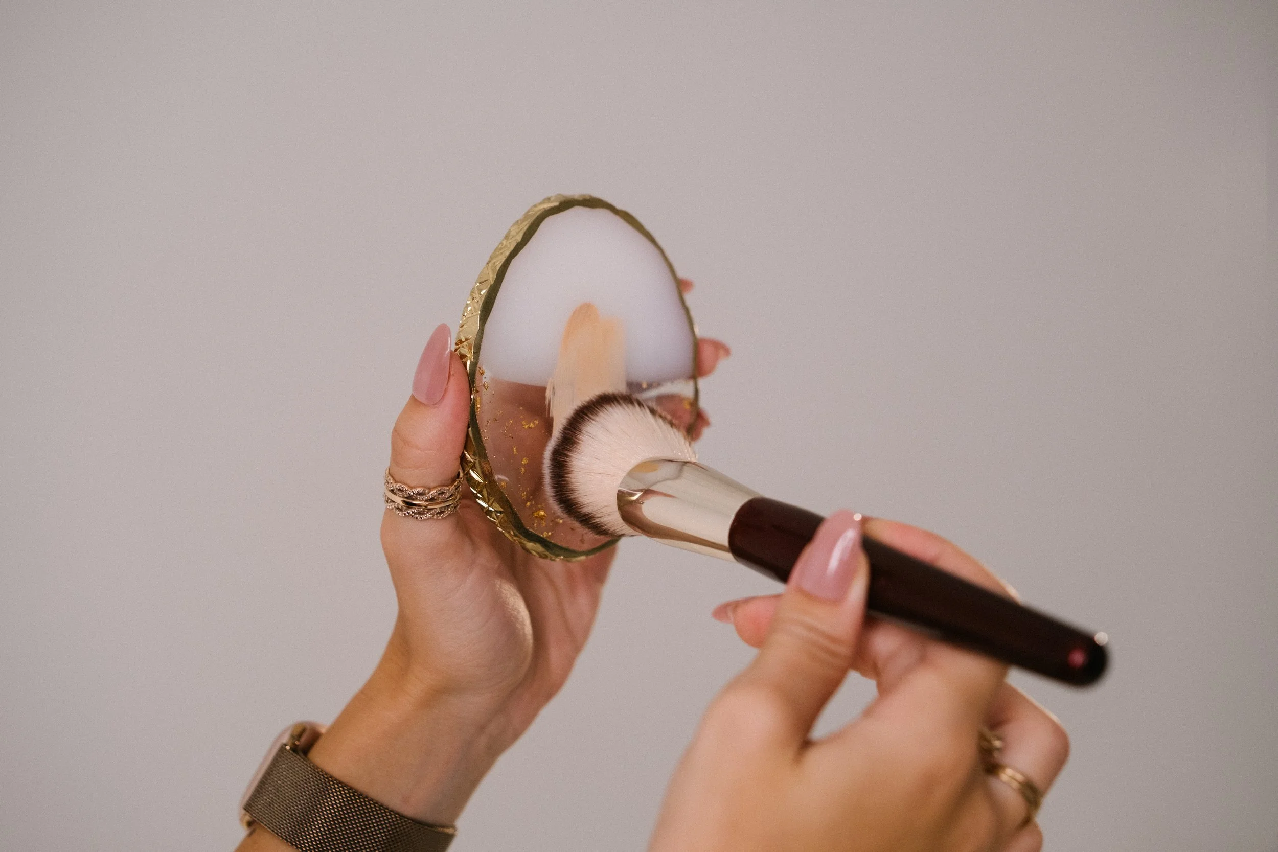 Person holding an oval-shaped mirror with gold trim and applying makeup with a brush.
