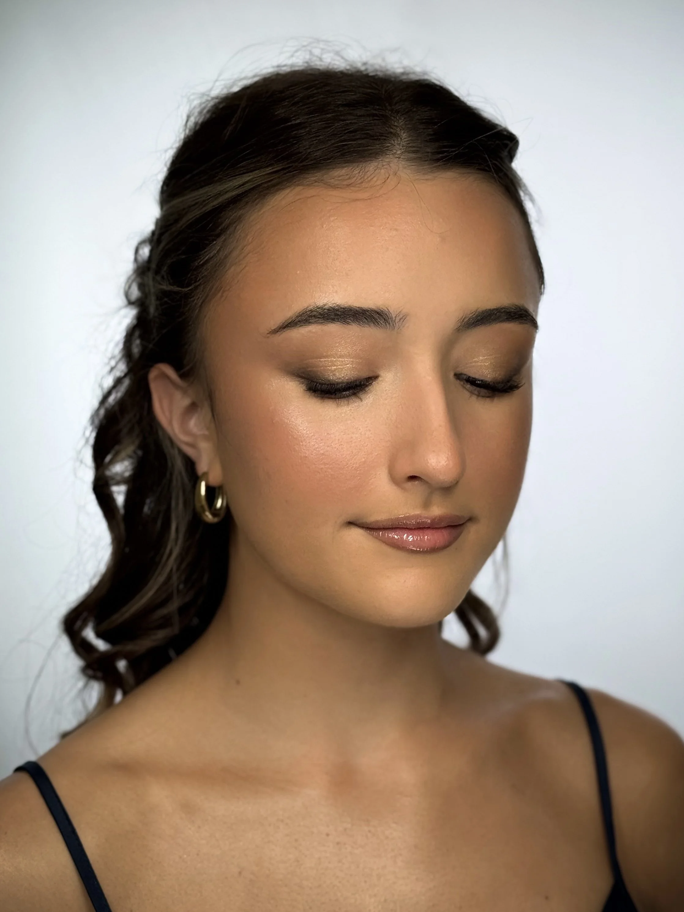 A woman with closed eyes, wearing makeup and gold hoop earrings, with dark wavy hair, against a plain background.
