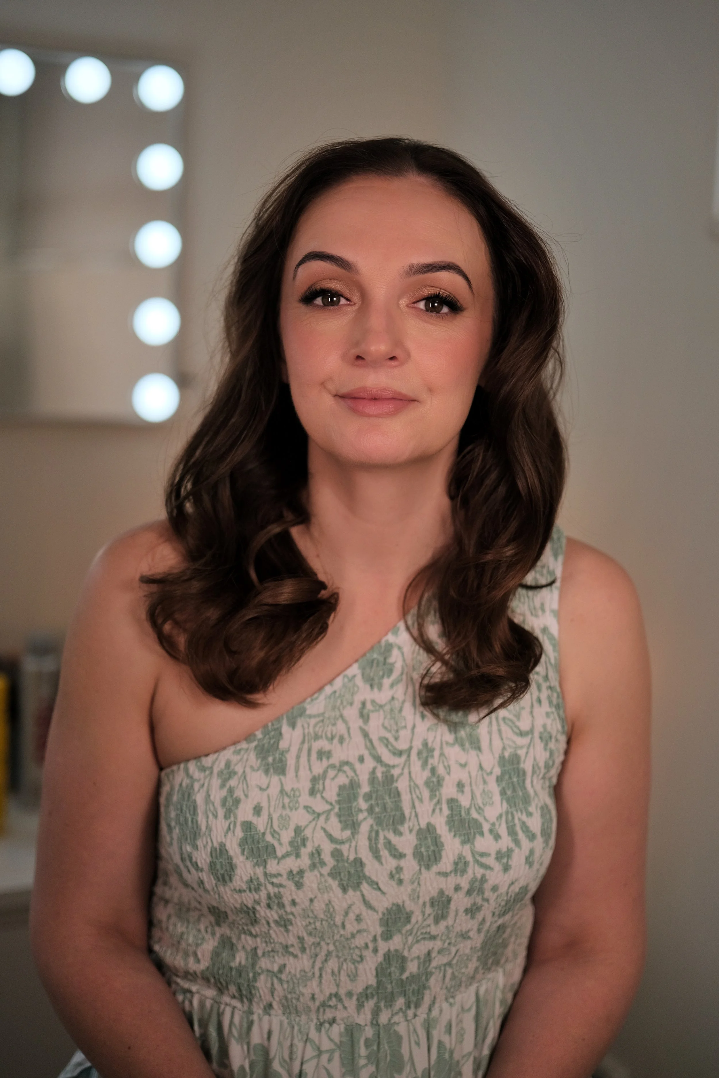 A woman with long, wavy brown hair and light makeup, wearing a sleeveless dress with green floral embroidery, standing in front of a mirror with bright lights.