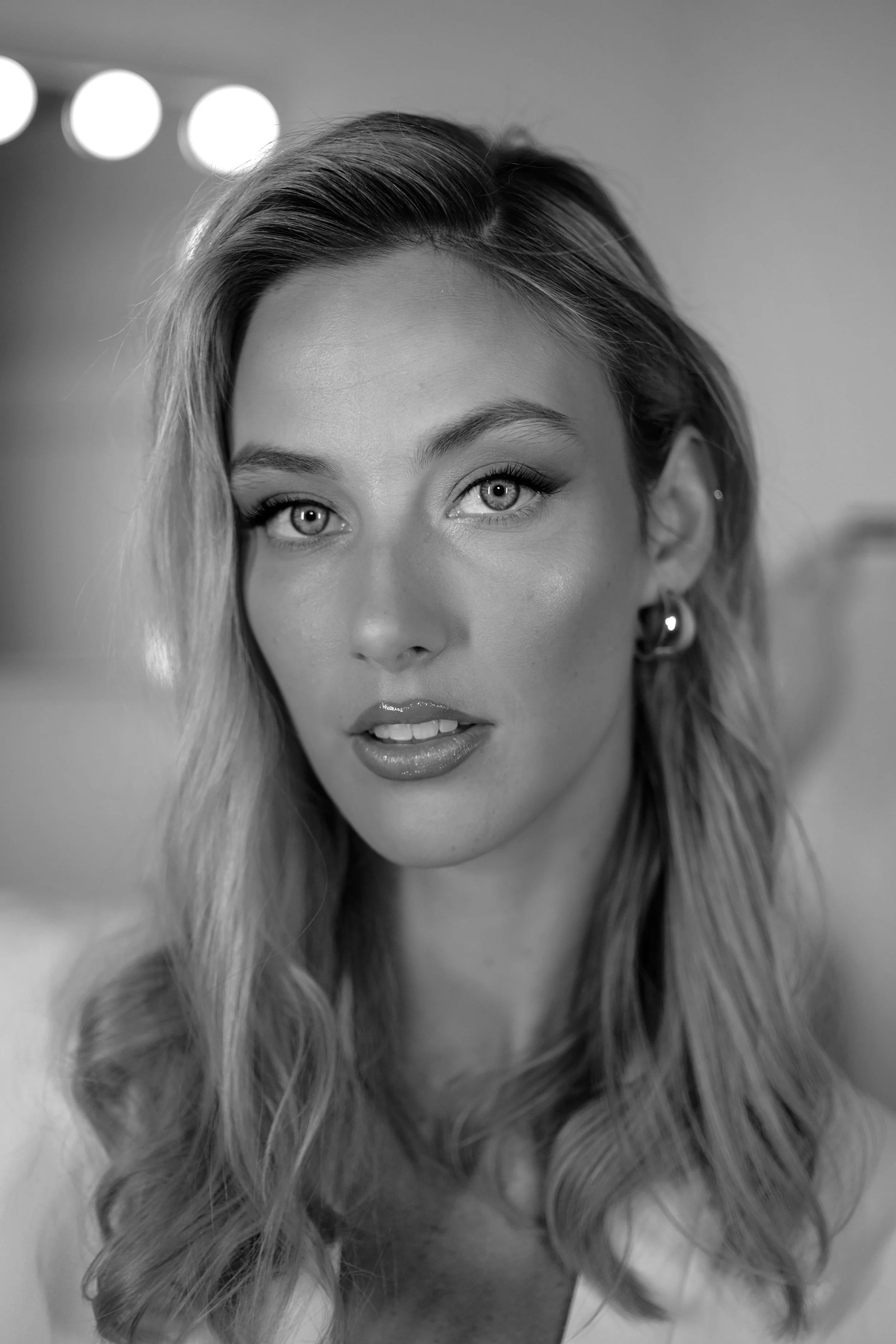 A black and white close-up portrait of a woman with wavy blonde hair, light-colored eyes, earrings, and makeup, looking directly at the camera.